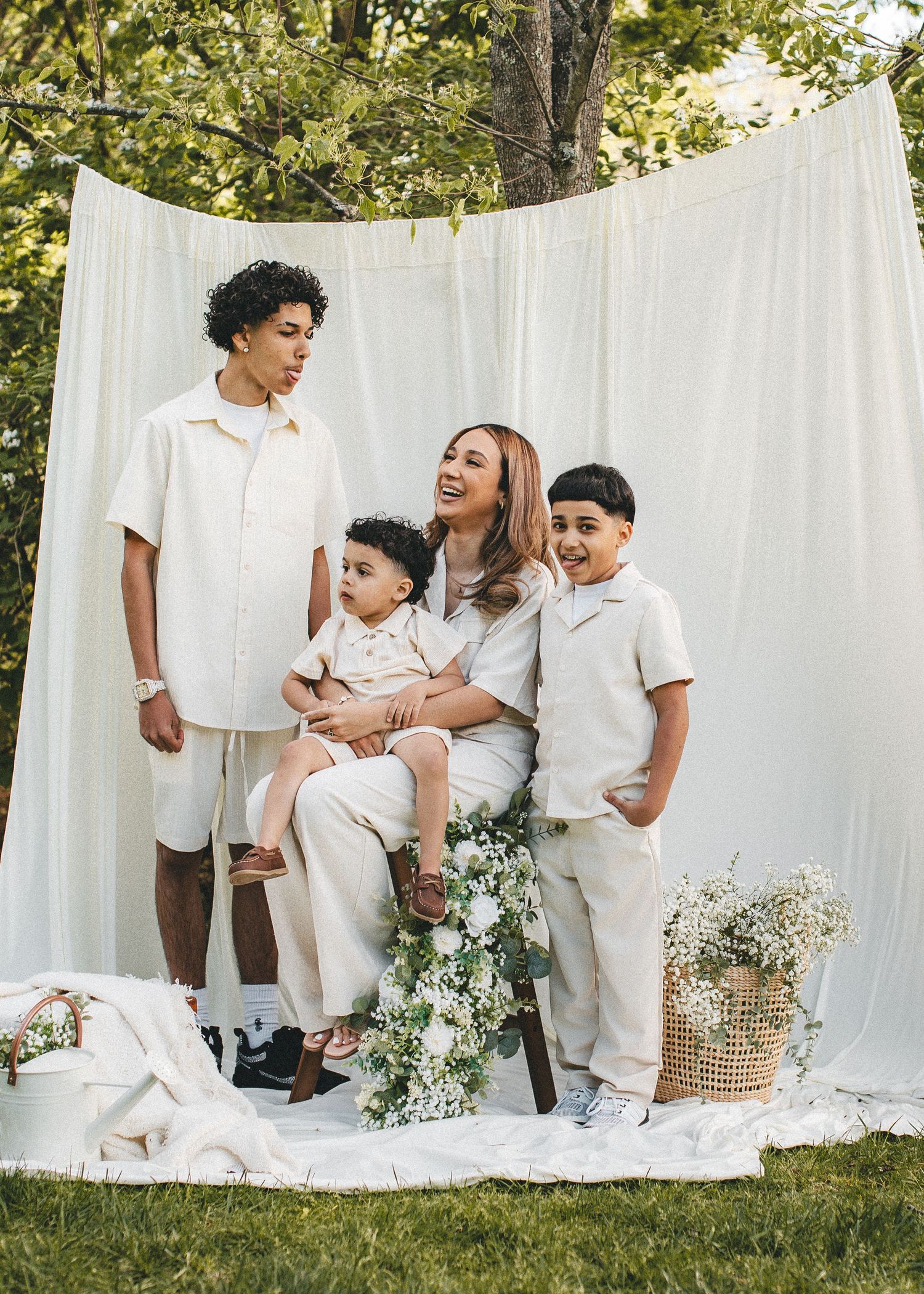 Mother seated outdoors with three sons in matching cream outfits against white drape backdrop.