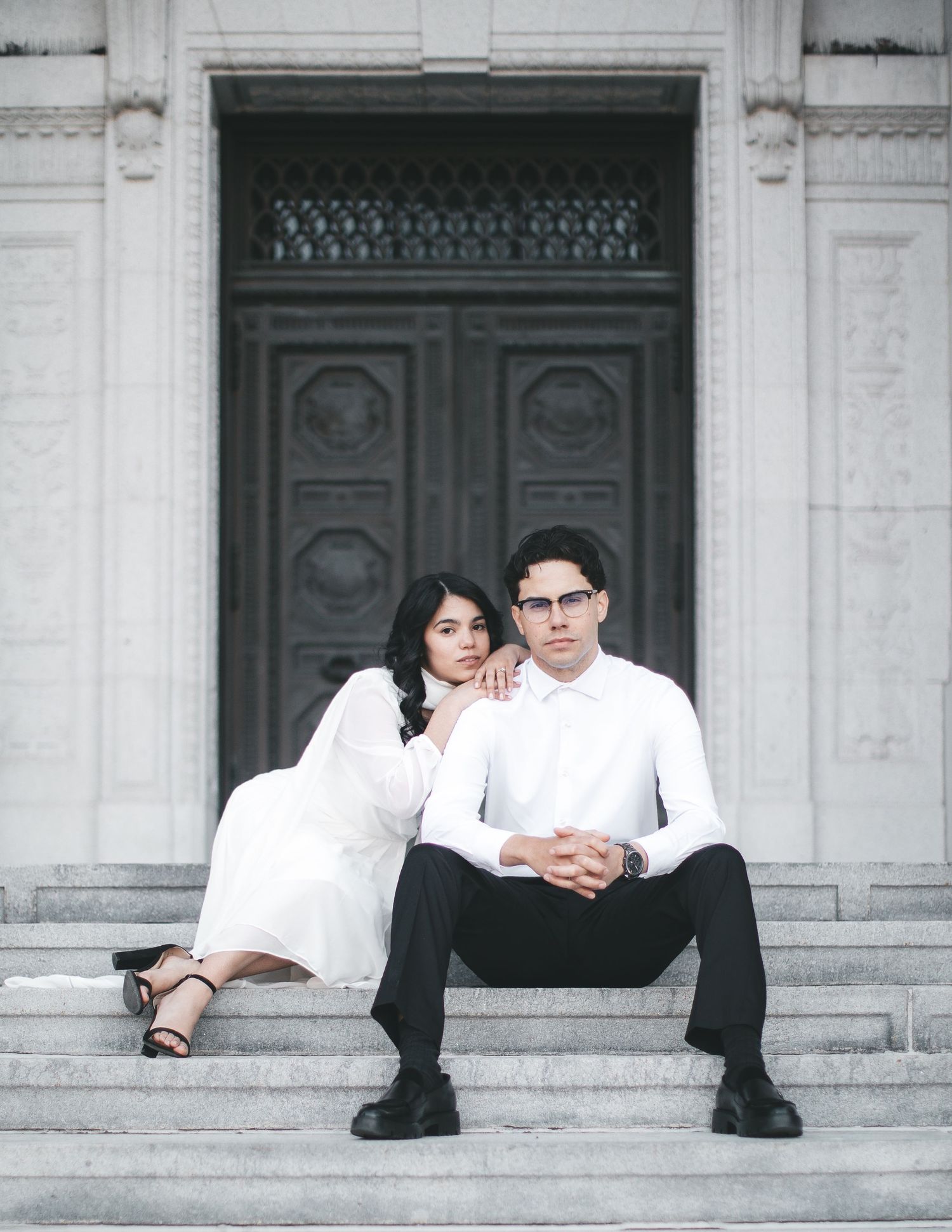 Elegant couple in white outfits sitting on stone steps before ornate doors for a formal portrait.