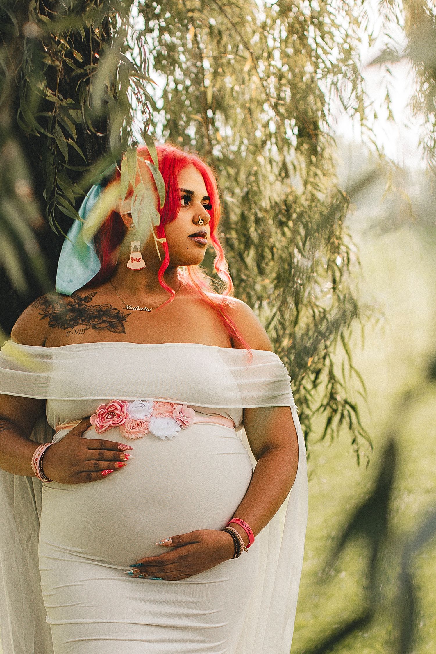 Pregnant woman in white off-shoulder gown with pink hair and floral accessories poses outdoors near willow tree.