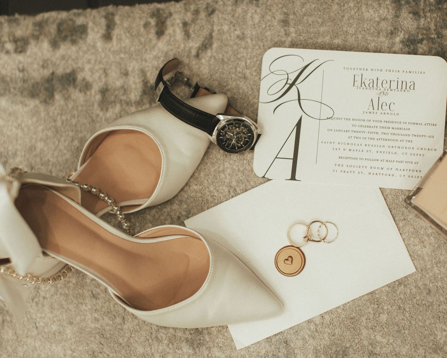 Wedding flat shoes, watch, rings, and personalized invitation arranged on a textured surface.