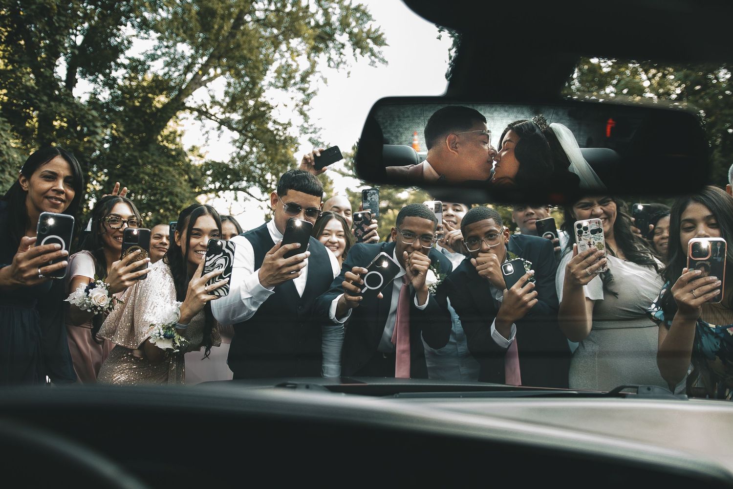 Couple kissing reflected in car mirror surrounded by wedding guests capturing the moment on smartphones.