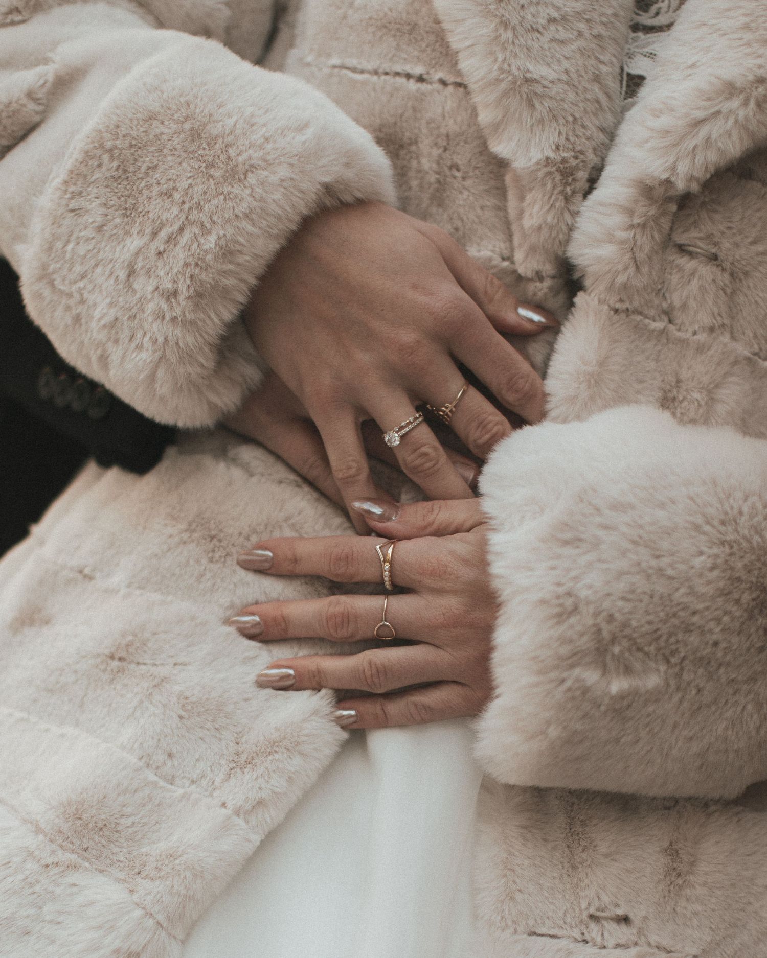 Close-up of bride's hands adorned with gold rings on a beige fur coat.