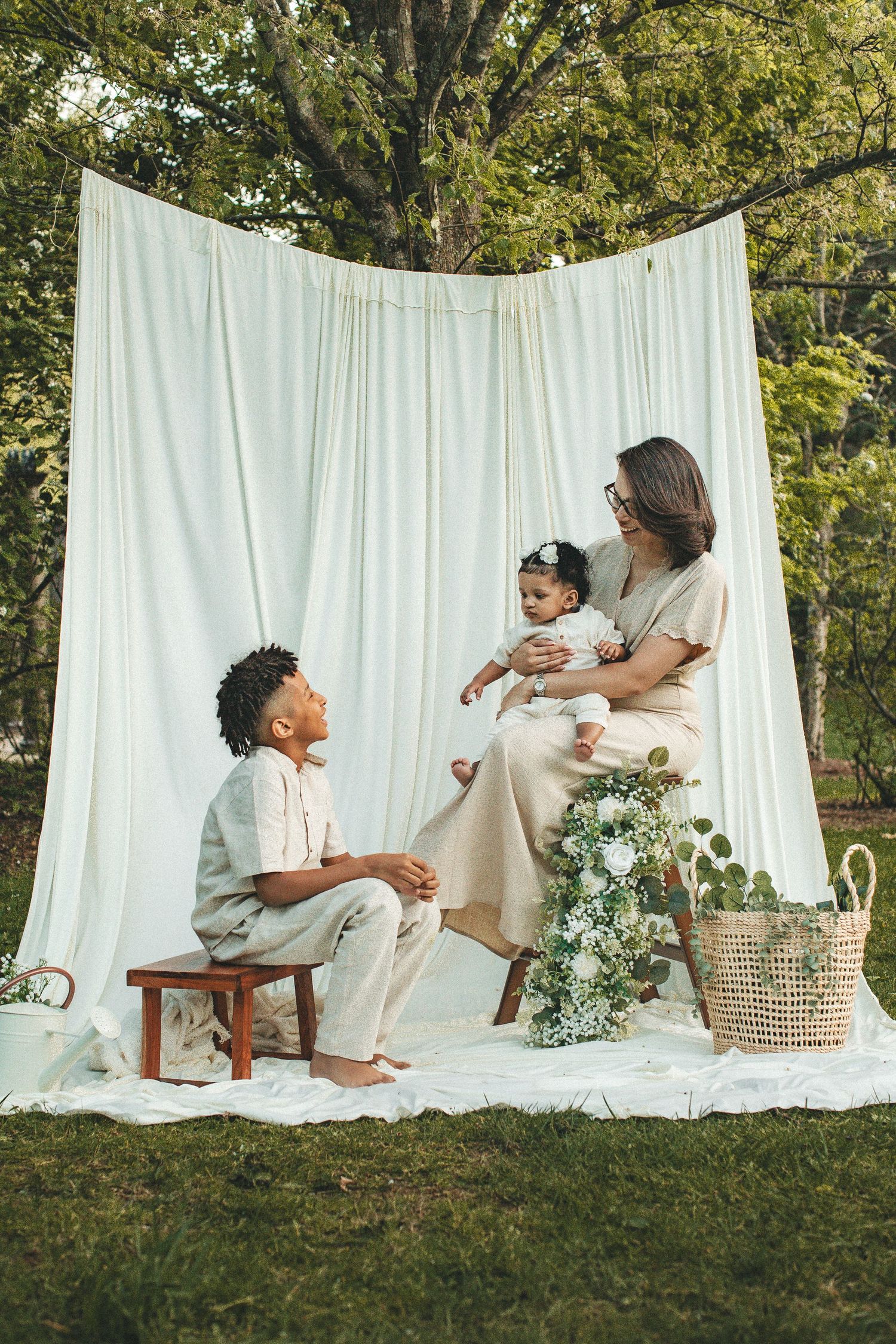 Family portrait outdoors with white backdrop, mother holding baby while son sits on stool.