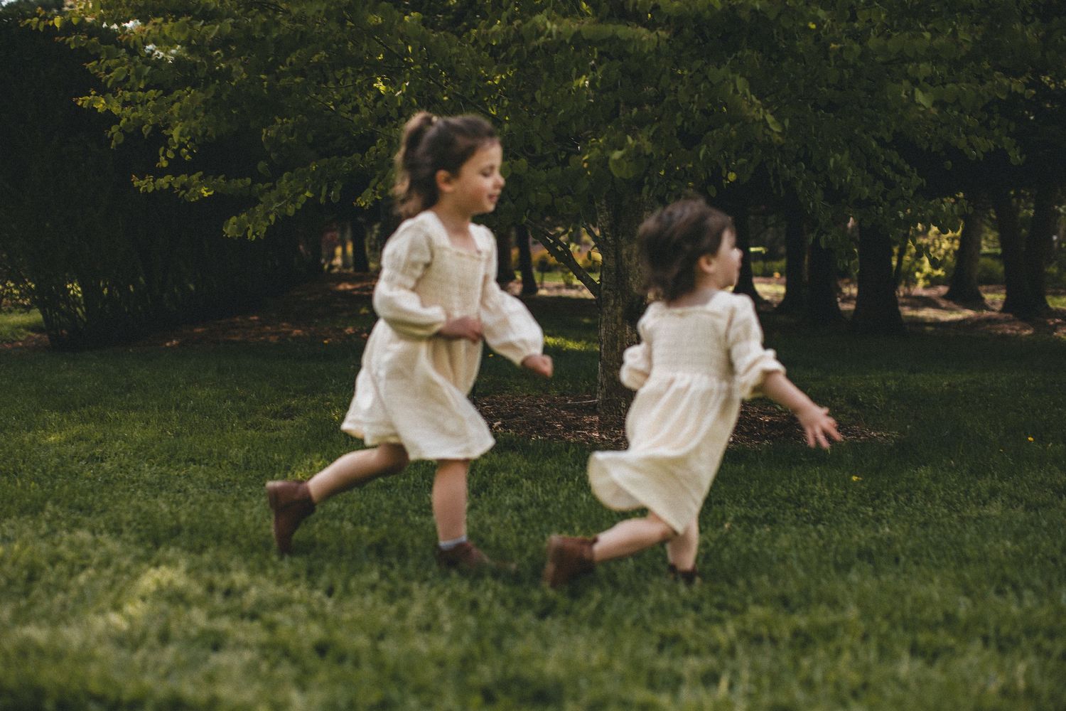 Two young girls in cream dresses holding hands and running on green grass in park.