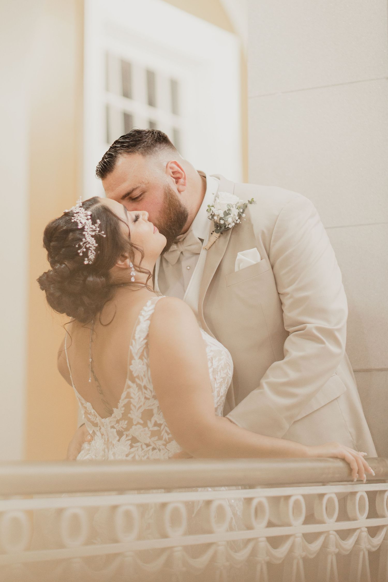 A bride and groom share a romantic kiss on a balcony, dressed in a white lace gown and beige suit.