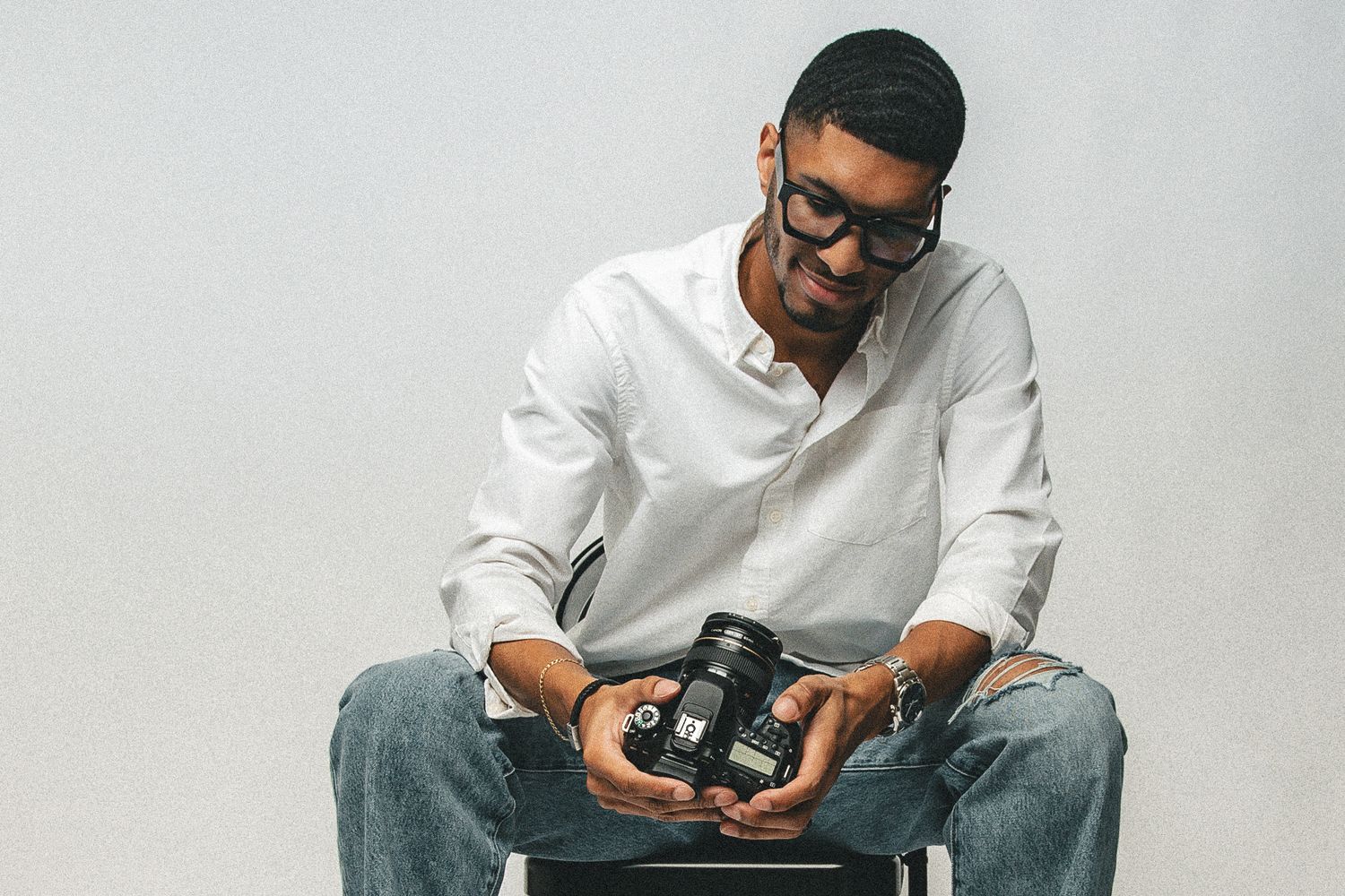 Young male photographer sitting and reviewing images on DSLR camera against light gray background.