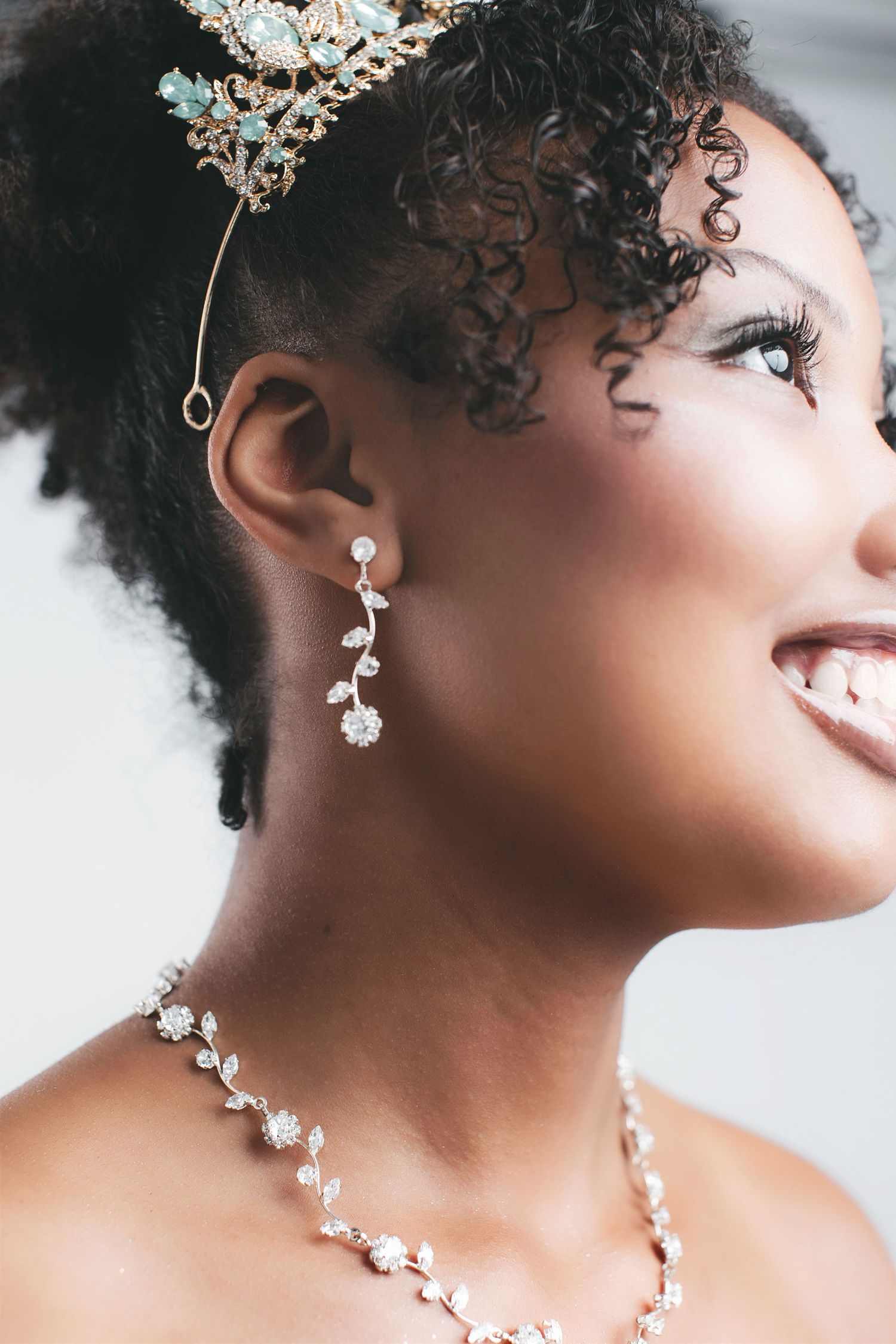 Smiling bride wearing crystal drop earrings, pearl necklace, and gold tiara with curly updo hairstyle.