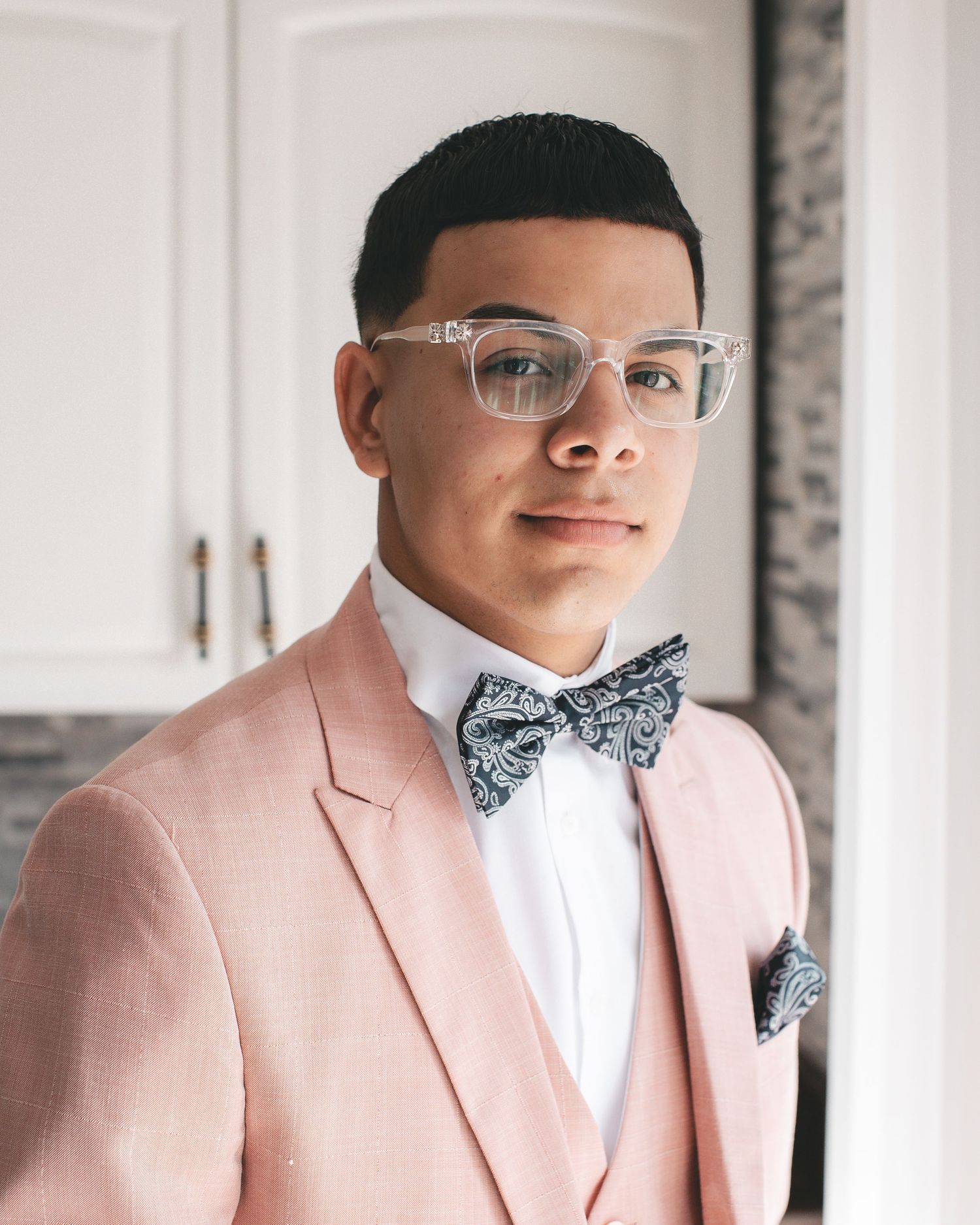 Young man in pink suit, white shirt, paisley bow tie, and clear glasses poses indoors for a formal occasion.