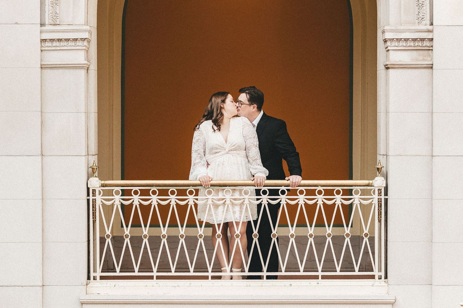 Couple sharing a romantic kiss on an ornate balcony with decorative iron railing at an elegant venue.