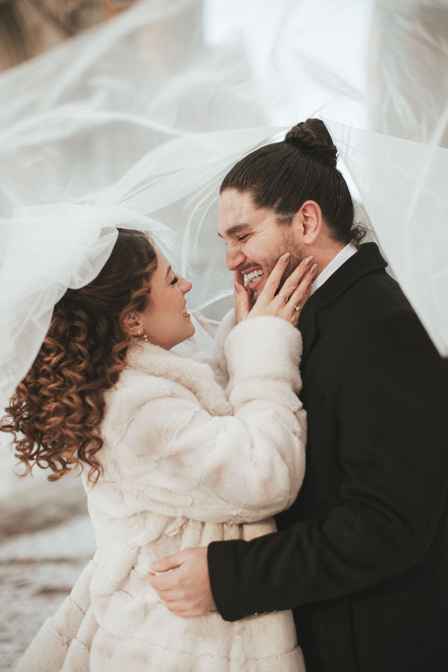 Smiling bride and groom sharing a tender moment under a flowing white veil in winter attire.