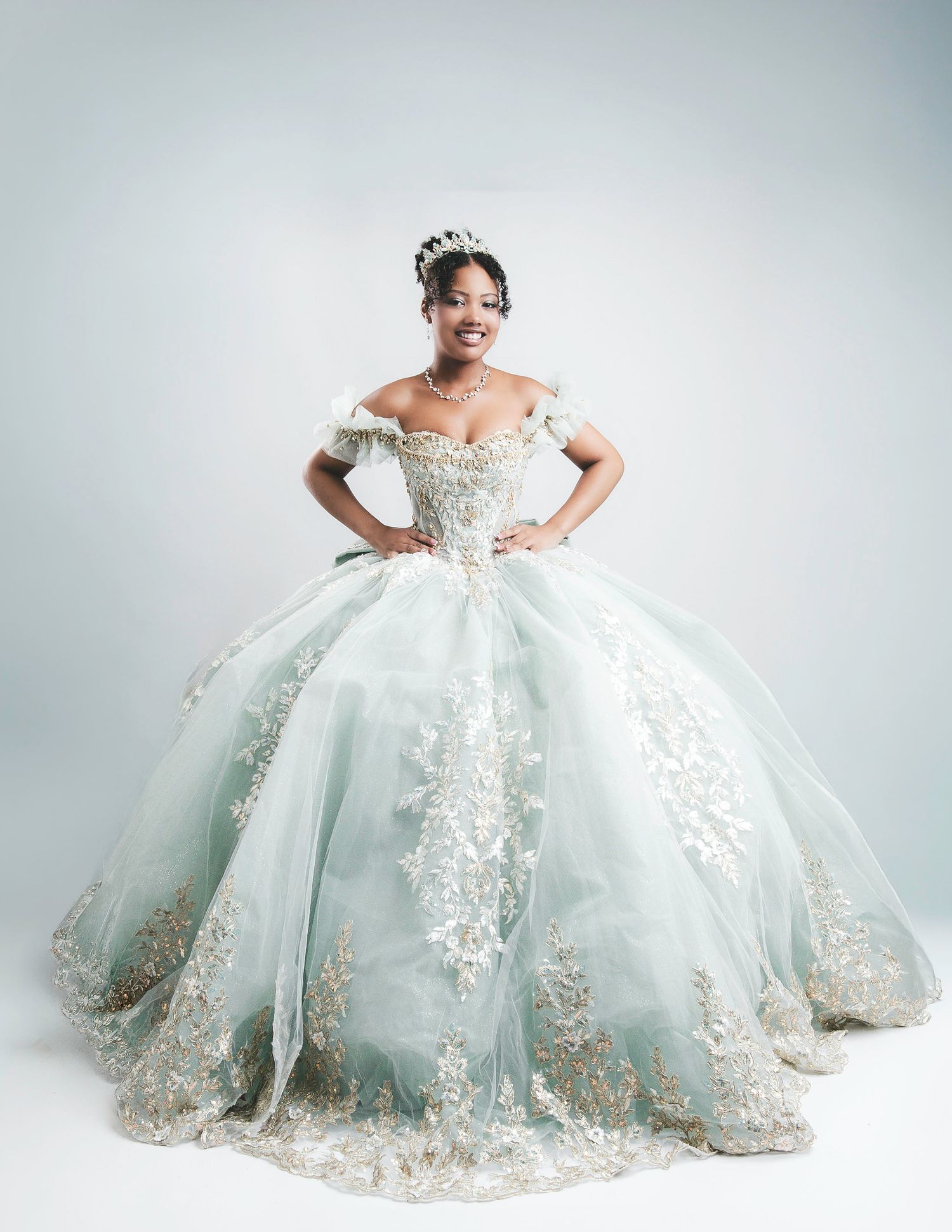 Young woman in mint green quinceañera ball gown with gold lace appliqués and tiara, posing confidently in studio.
