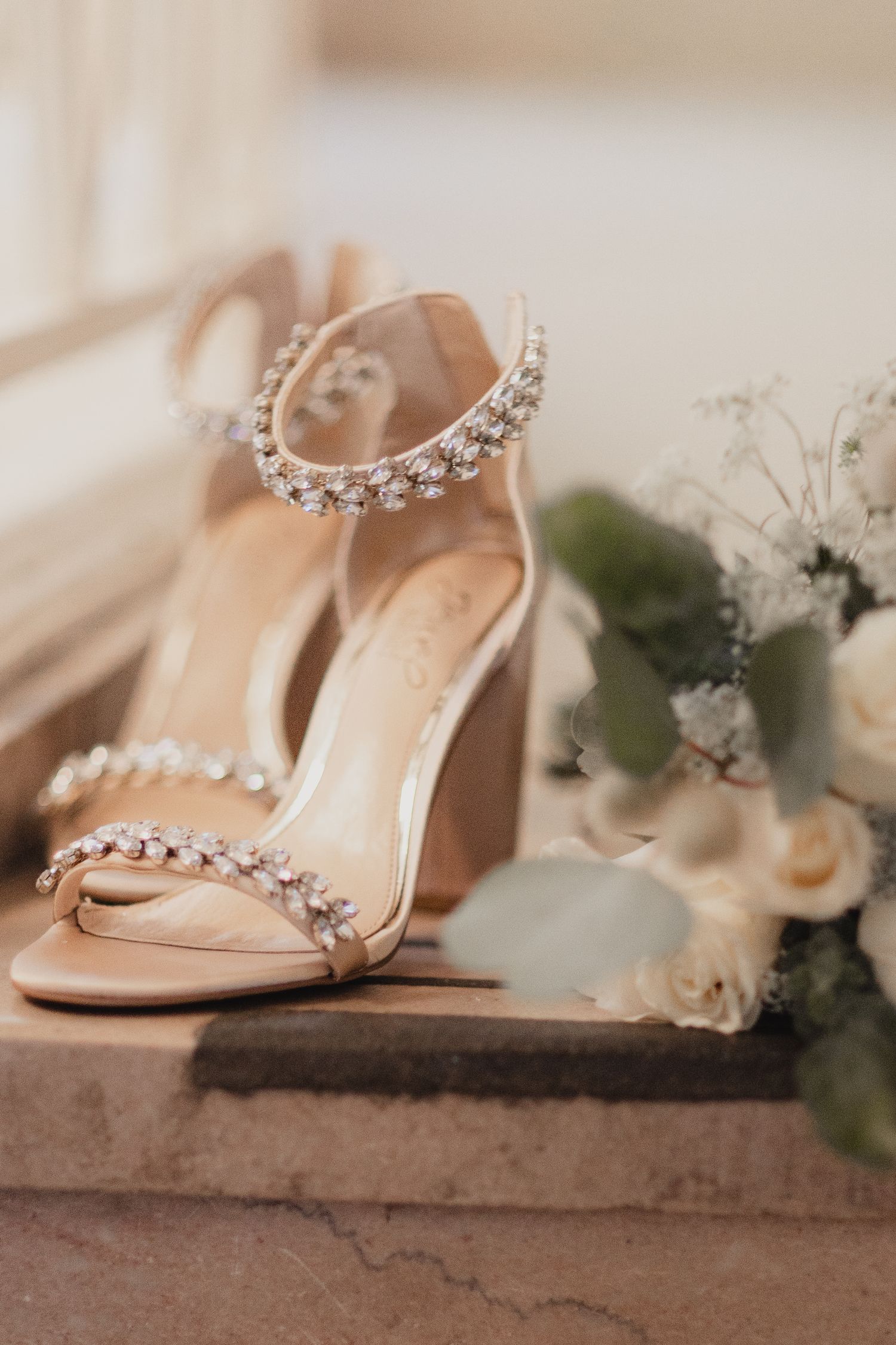 Elegant beige bridal heels adorned with crystal embellishments beside a white floral bouquet.