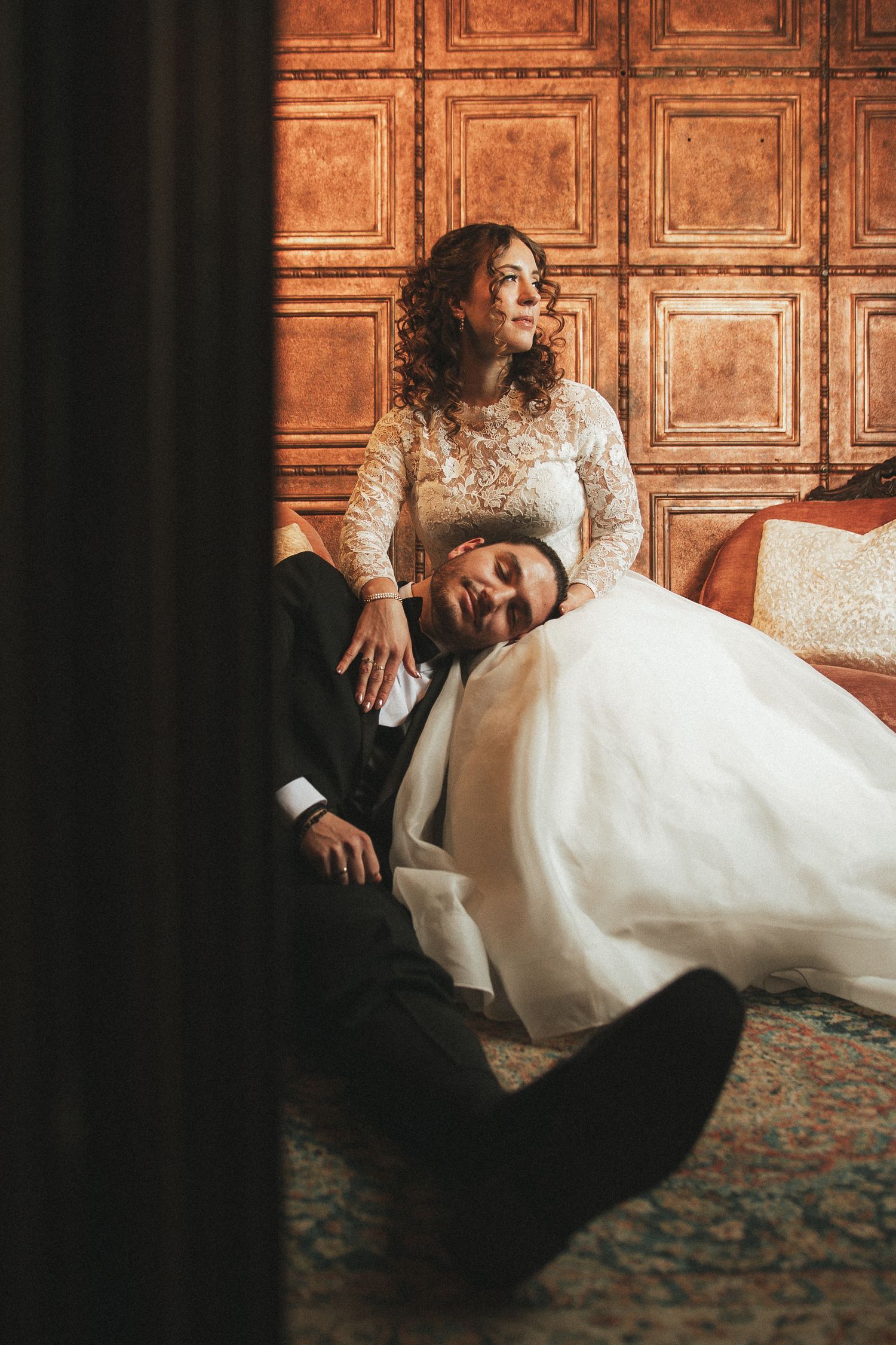 Bride in lace gown and groom in black tuxedo share intimate moment on floor of elegant wood-paneled room.