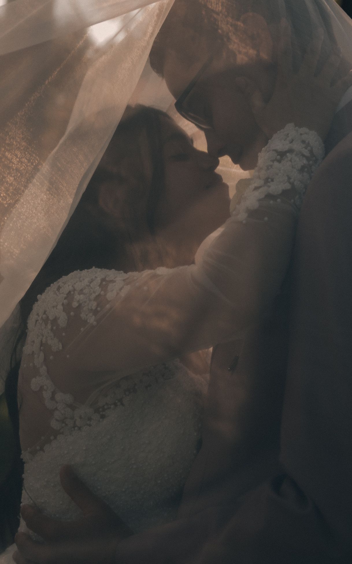 Romantic bride and groom sharing an intimate moment under a sheer wedding veil, captured in soft warm tones.