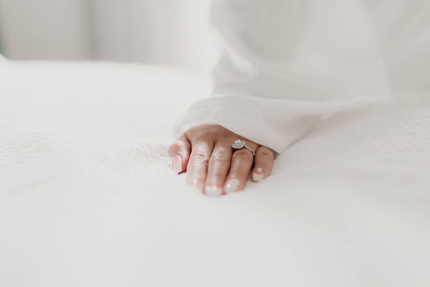 Close-up of a bride's hand wearing a diamond engagement ring, resting on a white surface in soft natural light.