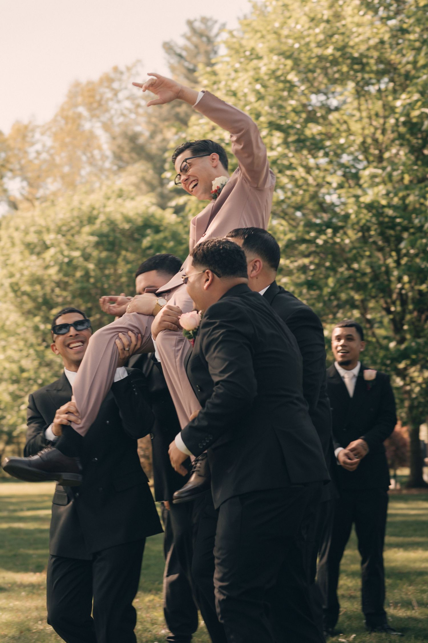 Groomsmen lifting groom in the air outdoors, celebrating at a wedding in a lush green park setting.