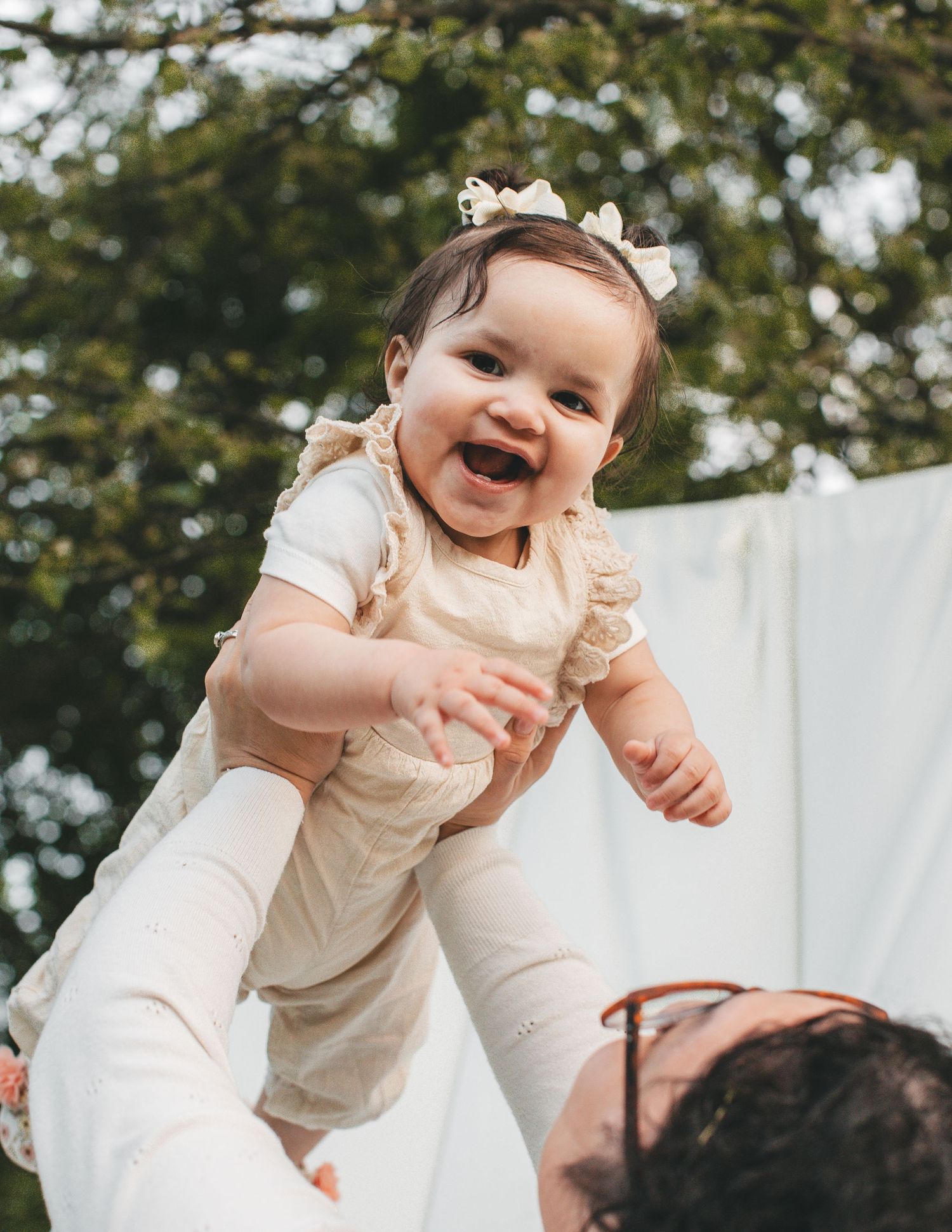 Joyful baby girl in cream outfit lifted in the air, laughing with trees in background.