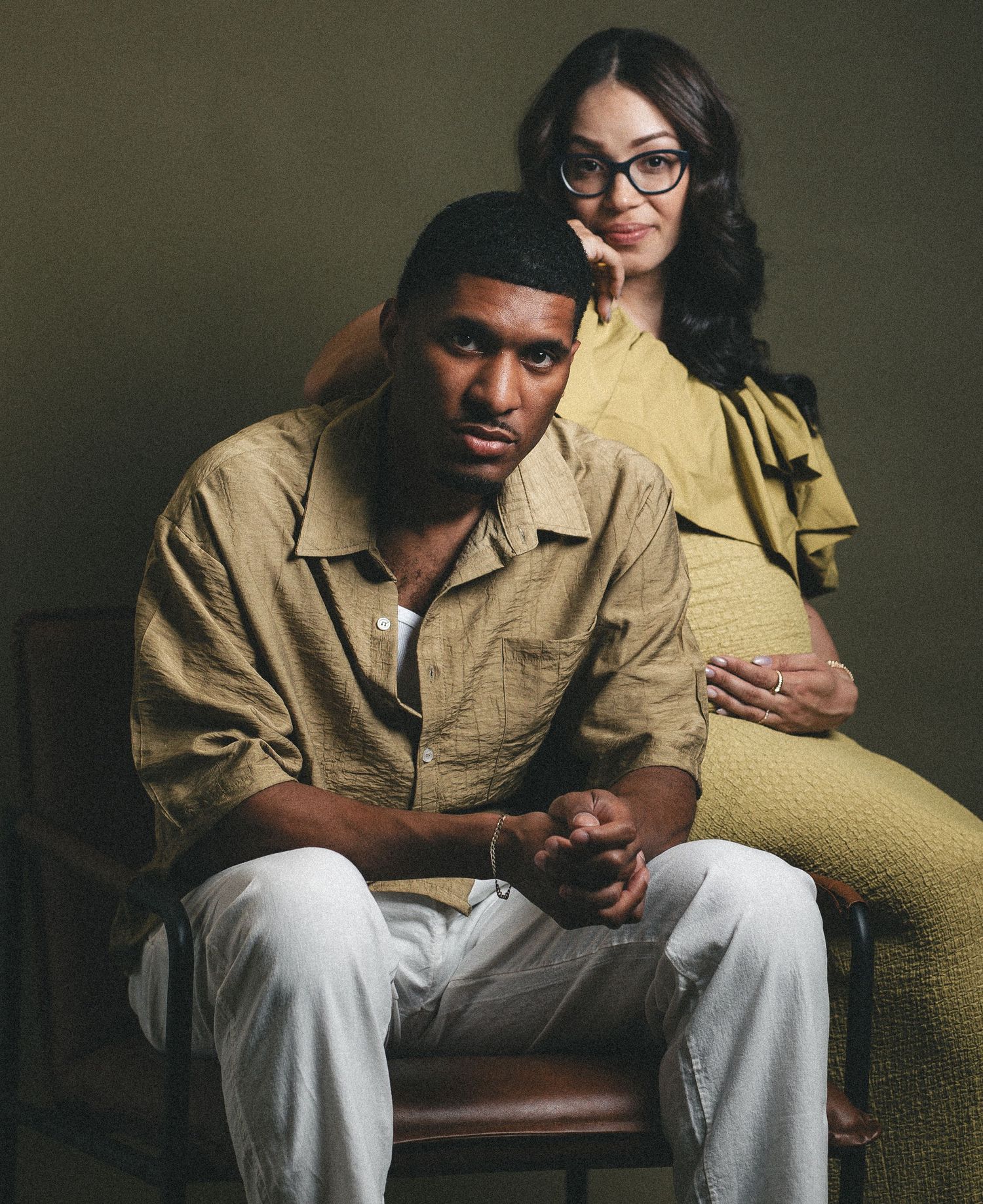 Studio portrait of pregnant couple, man seated in chair with woman standing behind him.