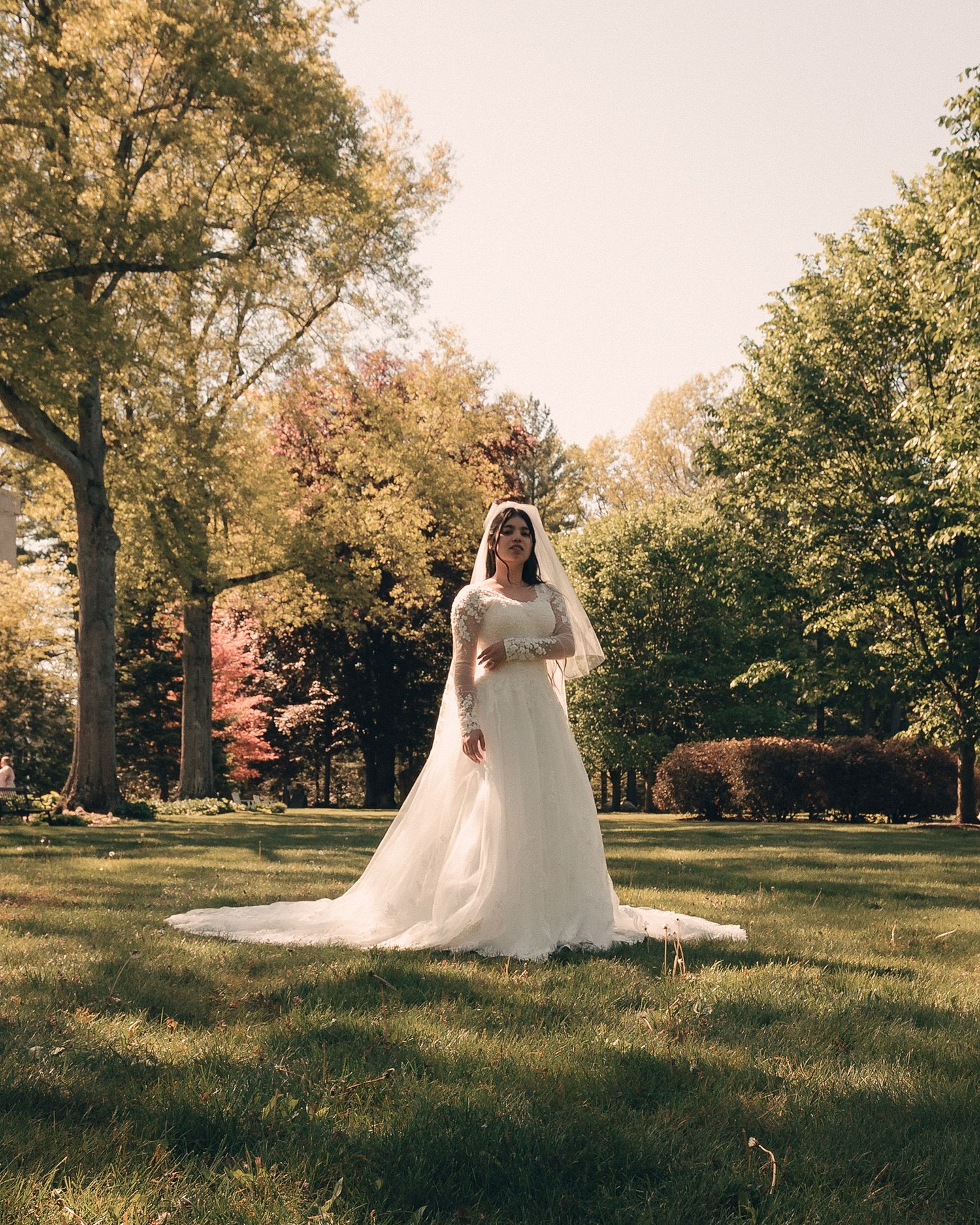 Bride in white wedding gown and veil standing on lush green lawn surrounded by autumn trees in sunlit park.