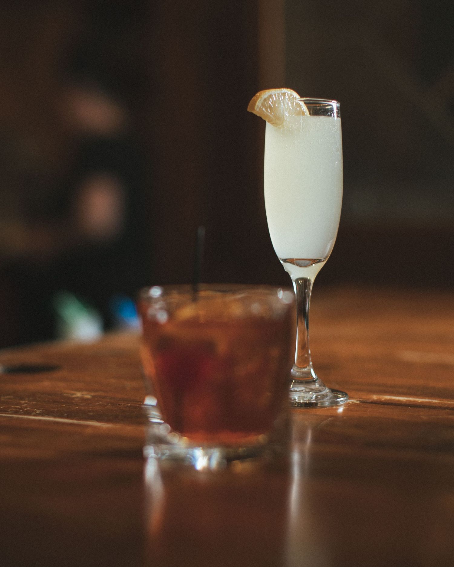Two cocktails on wooden bar, a dark whiskey and a frothy champagne flute with lemon.
