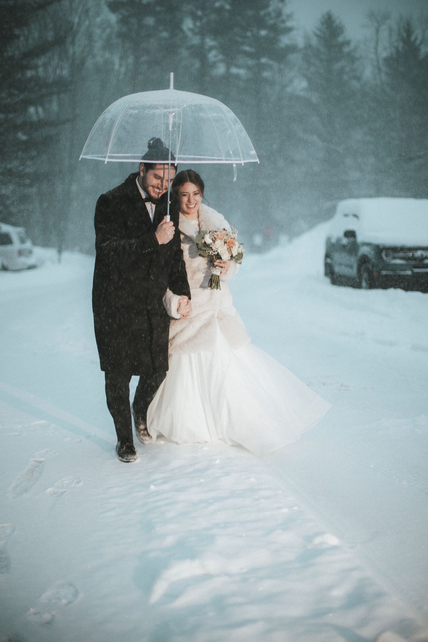 Bride and groom walking under clear umbrella in heavy snowstorm, bride holding bouquet in white gown.