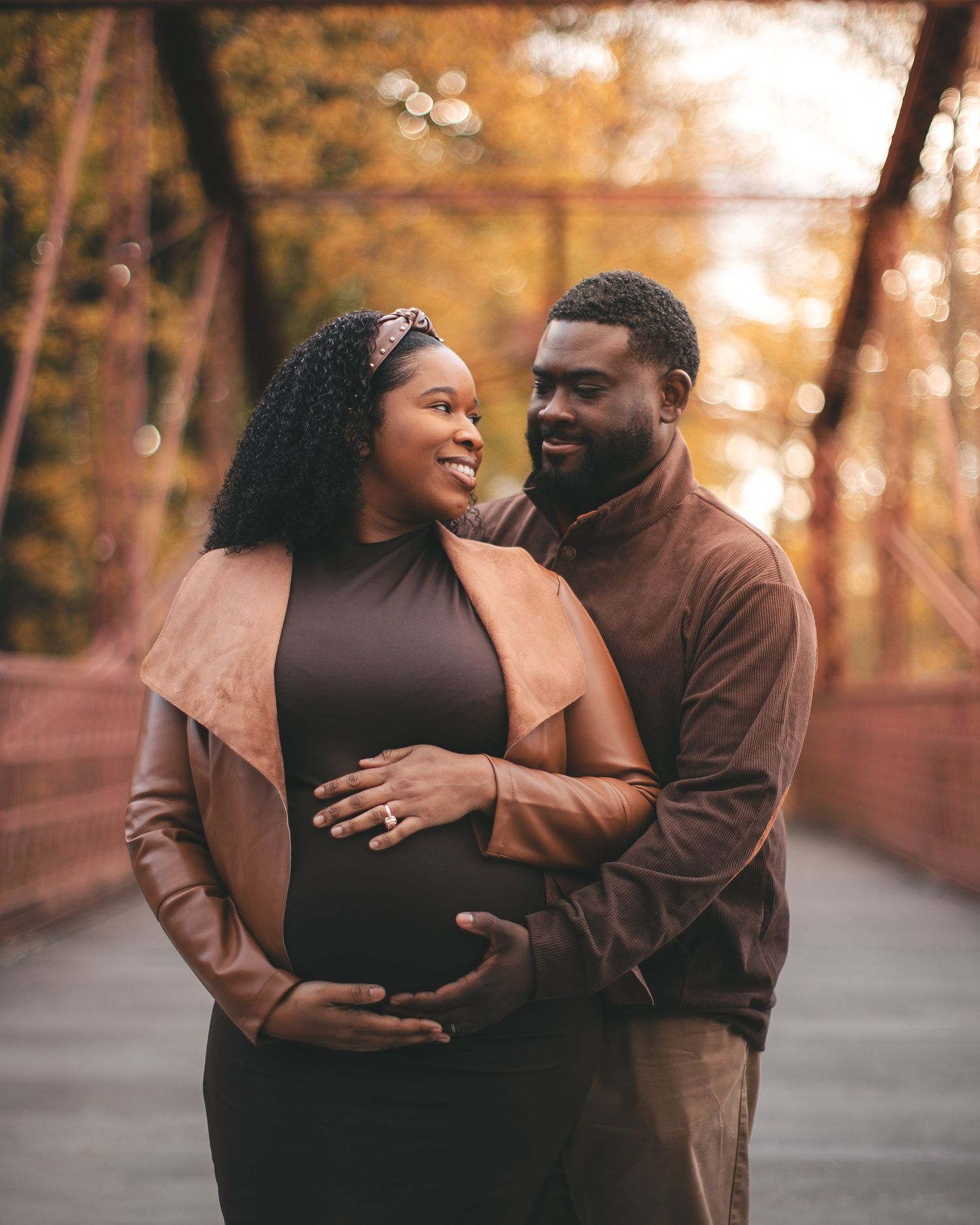 A pregnant Black couple poses on a bridge surrounded by autumn foliage, both smiling and cradling her baby bump.