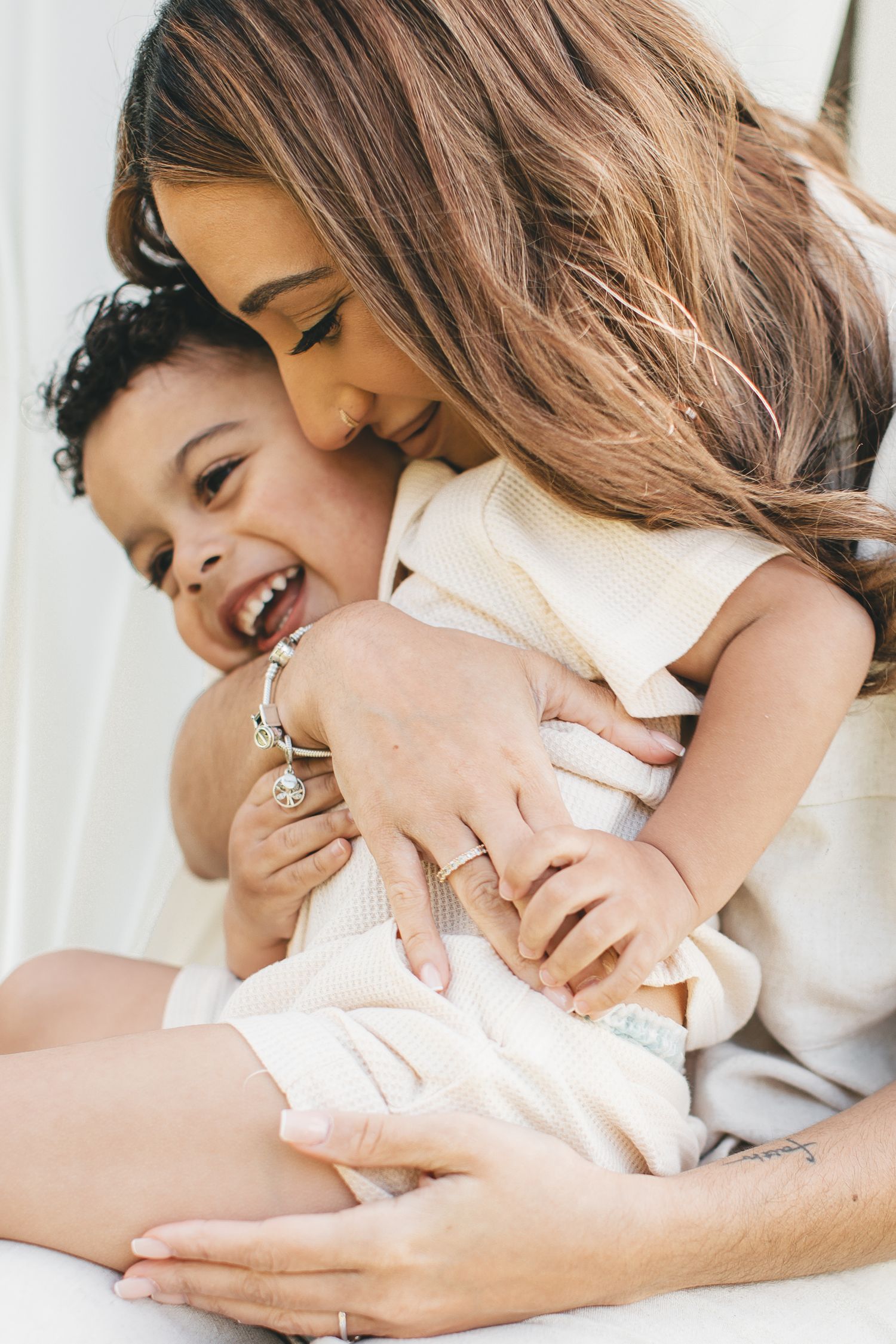 Woman with long hair hugging laughing toddler while holding newborn baby in white setting.