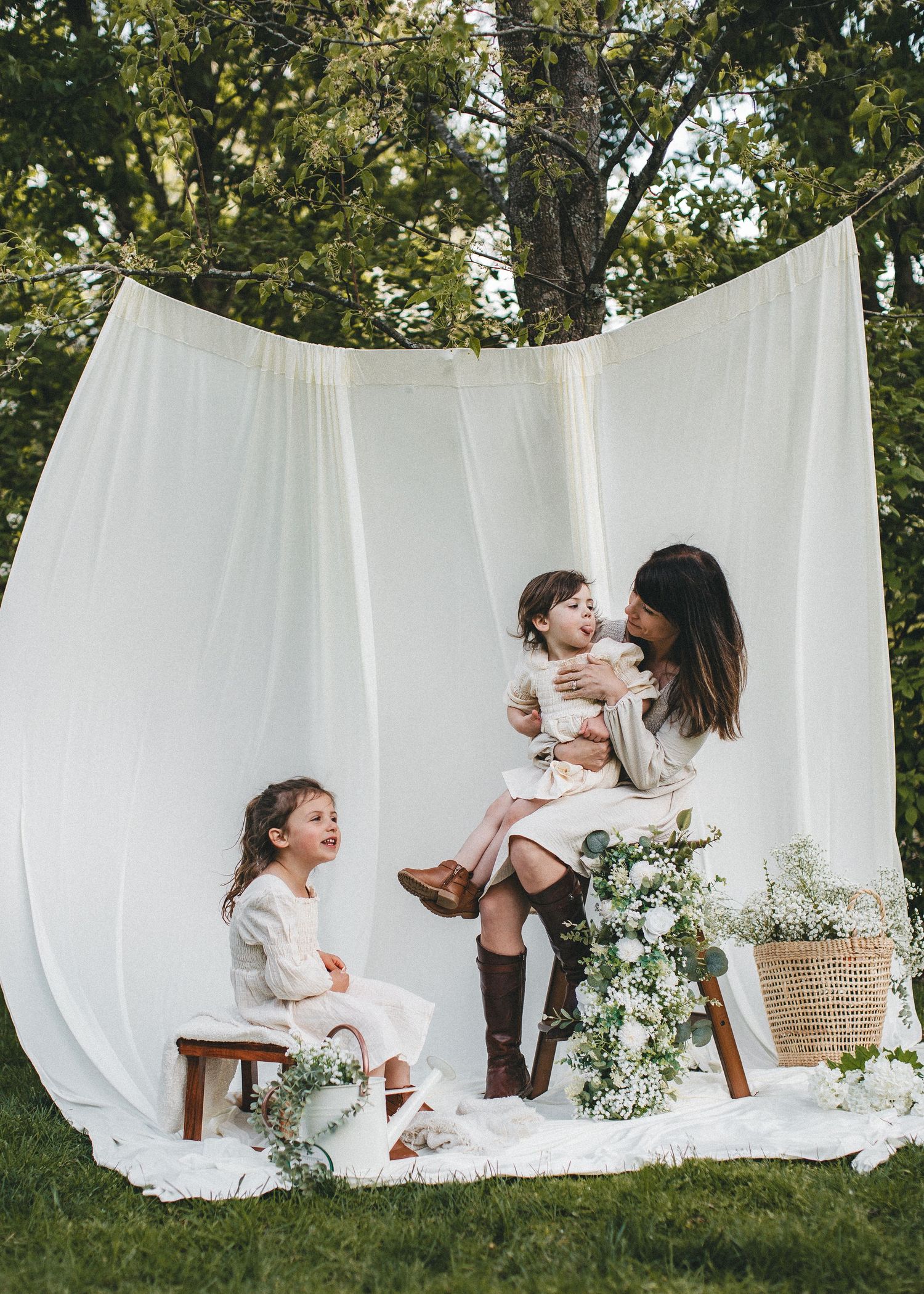 Mother holds toddler while young girl sits on stool, posing outdoors with white fabric backdrop and floral arrangements.