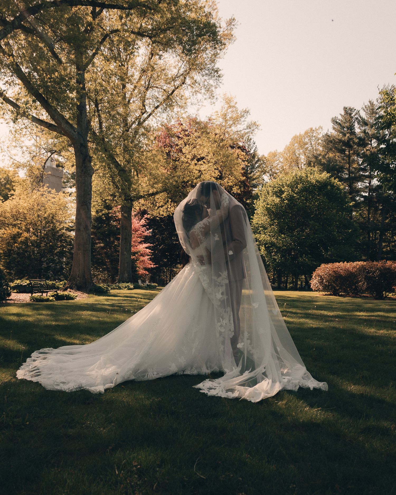Bride in a flowing white gown and long veil standing in a lush green garden surrounded by trees.