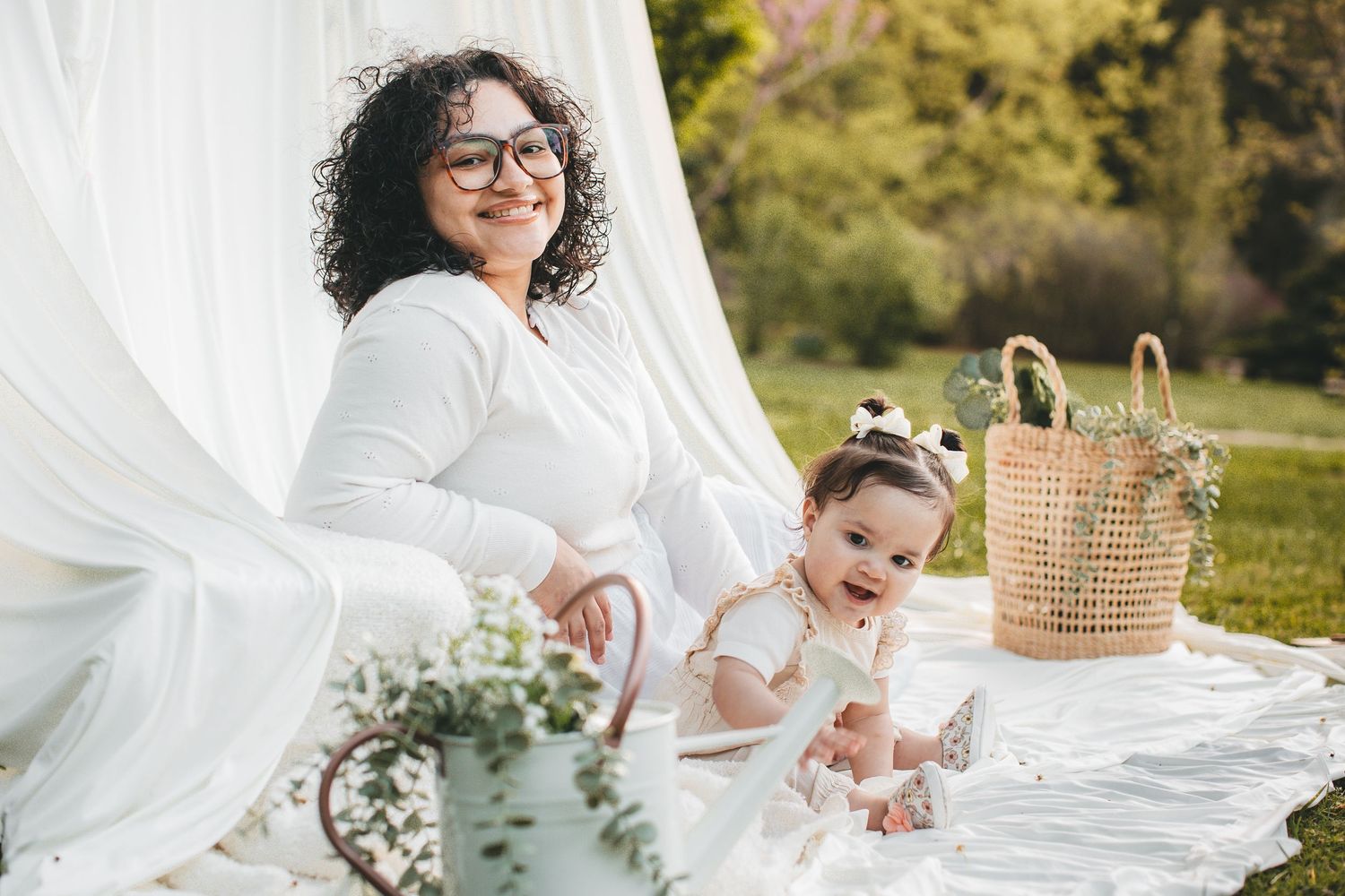 Mother with curly hair and glasses sitting outdoors with baby on white blanket backdrop.
