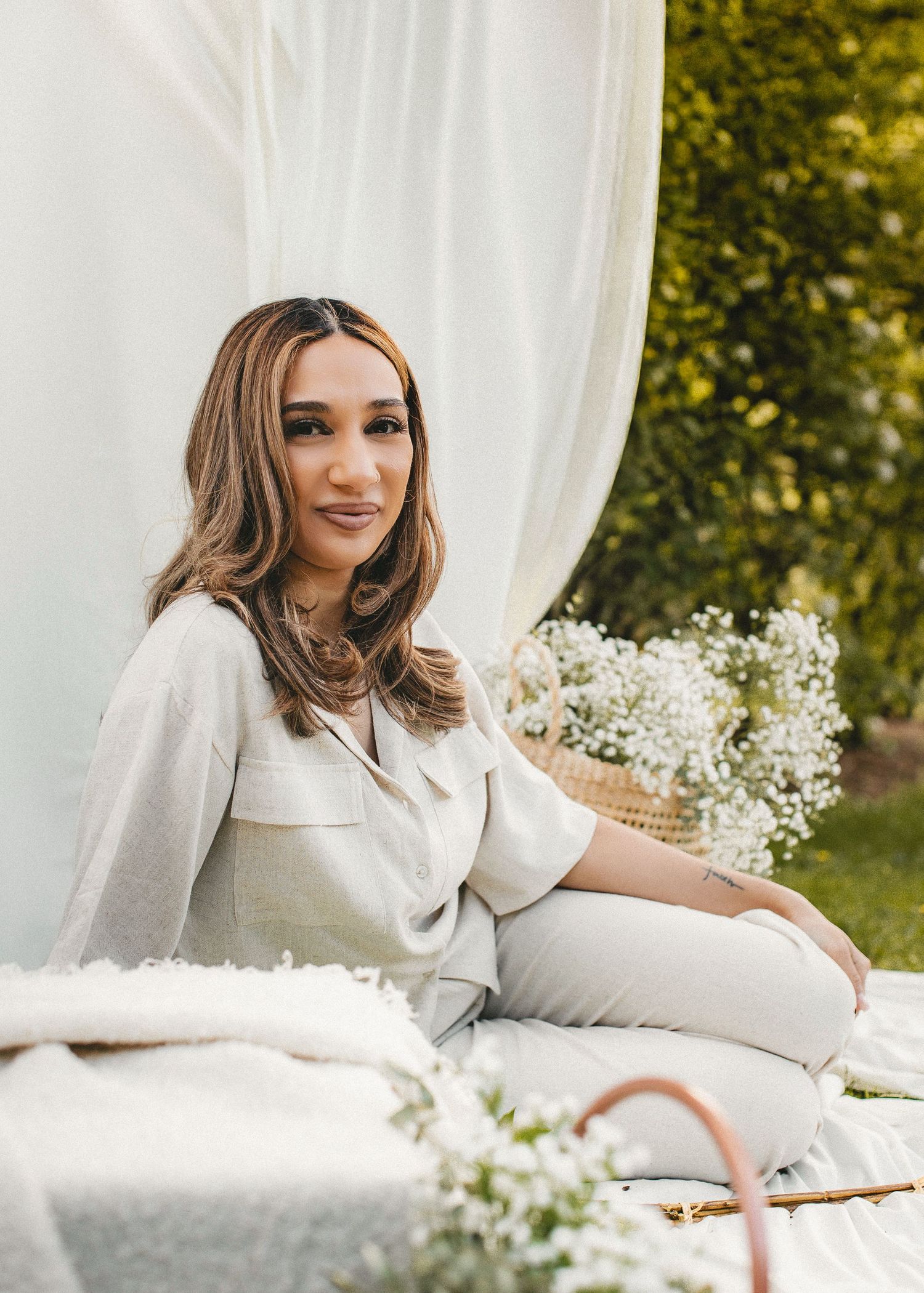 Woman with long brown hair sitting outdoors on white blanket surrounded by white flowers.