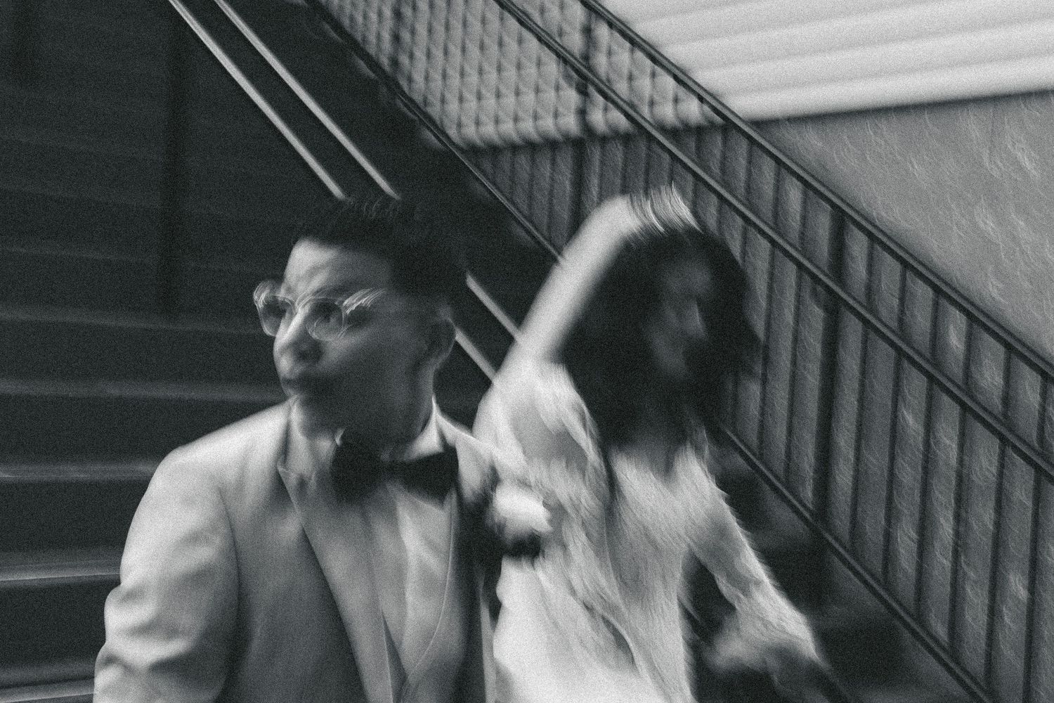 Black and white photo of a couple in formal attire walking near staircase, motion blur effect.