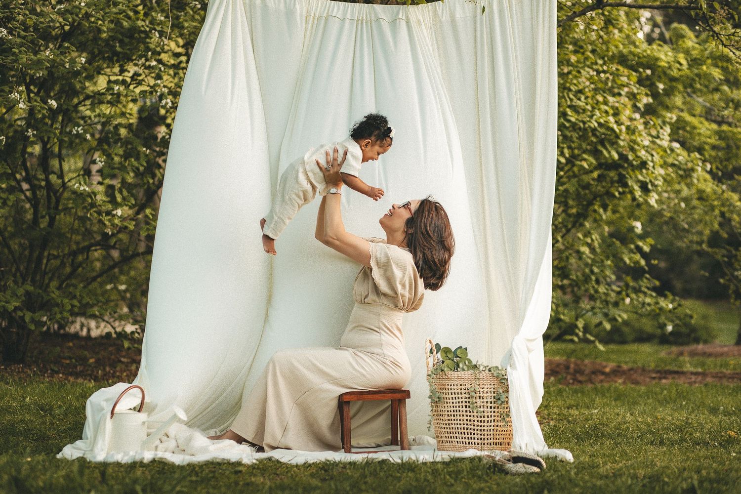 Mother lifting smiling baby in the air outdoors with white drape backdrop and green trees.
