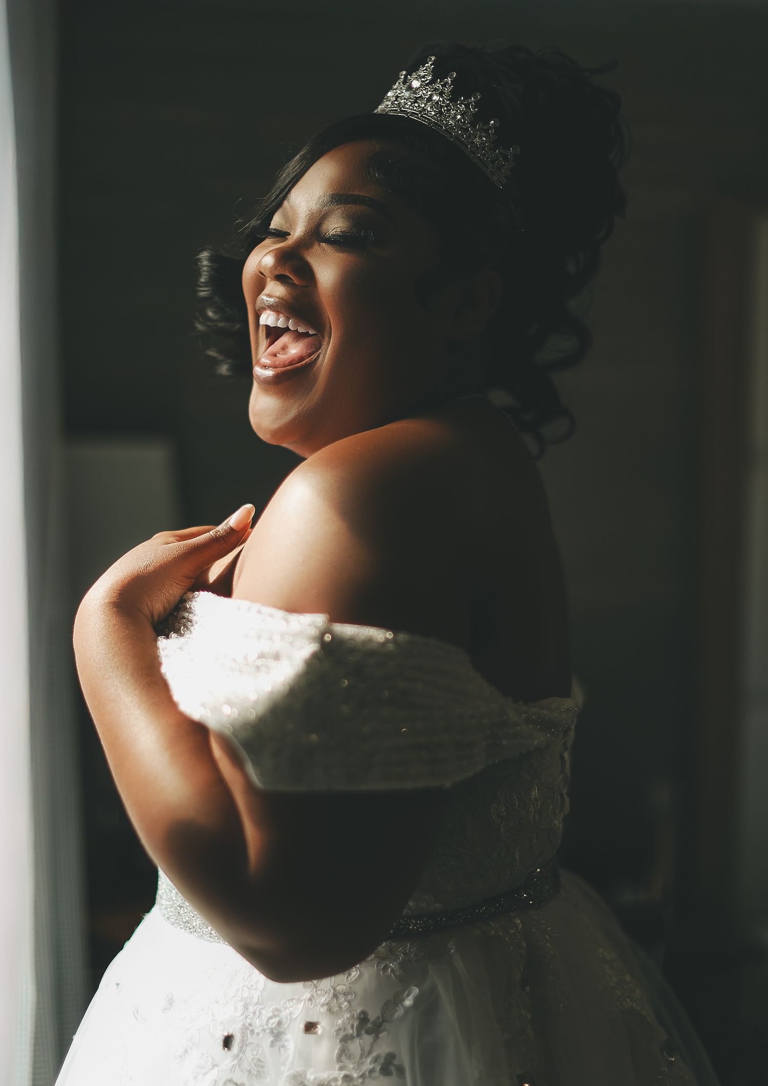Joyful bride in white off-shoulder gown and tiara laughing radiantly near window light on wedding day.