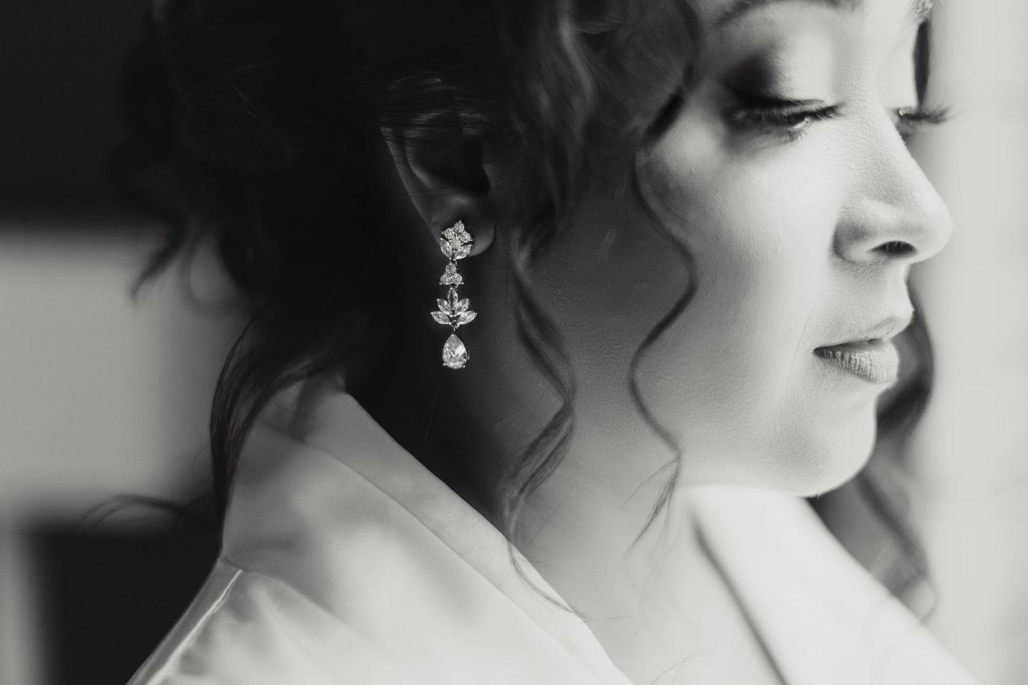 Black and white close-up of a bride wearing elegant crystal drop earrings, with soft curled hair and a white robe.