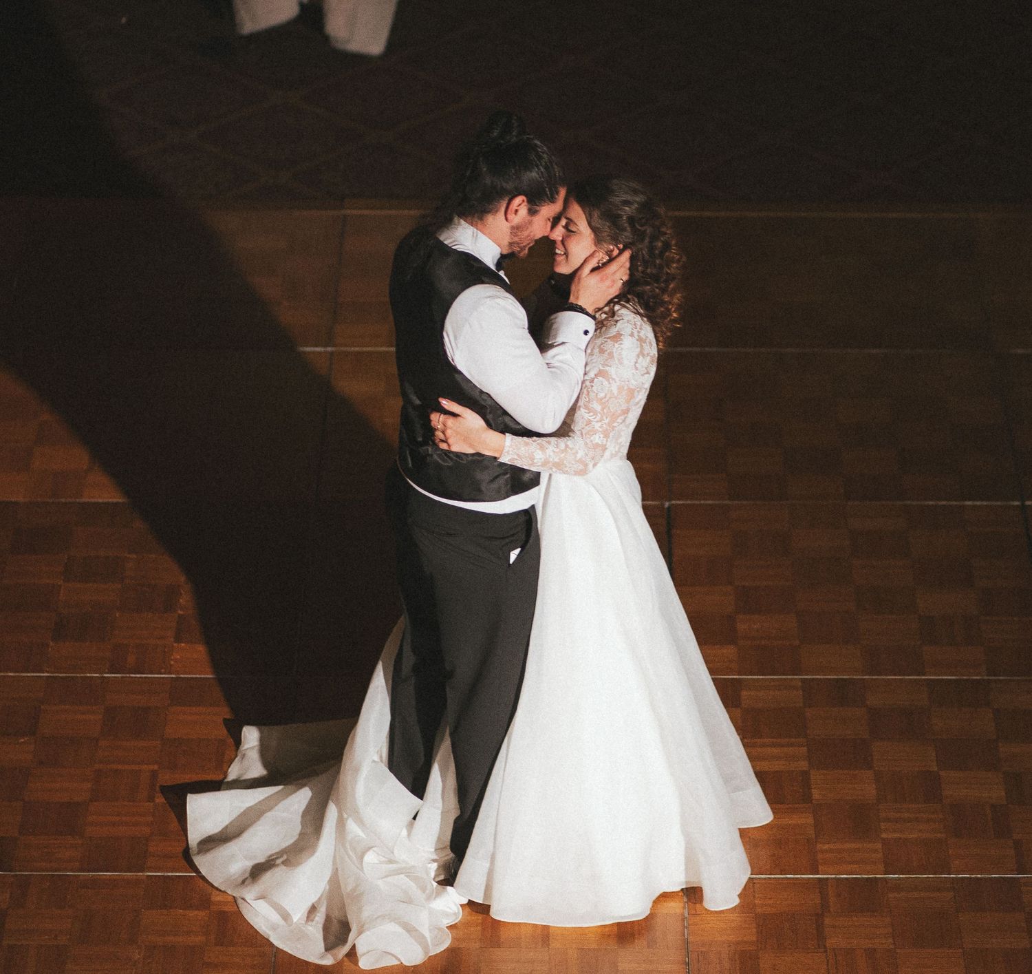 Bride and groom sharing first dance in wedding attire on wooden dance floor.