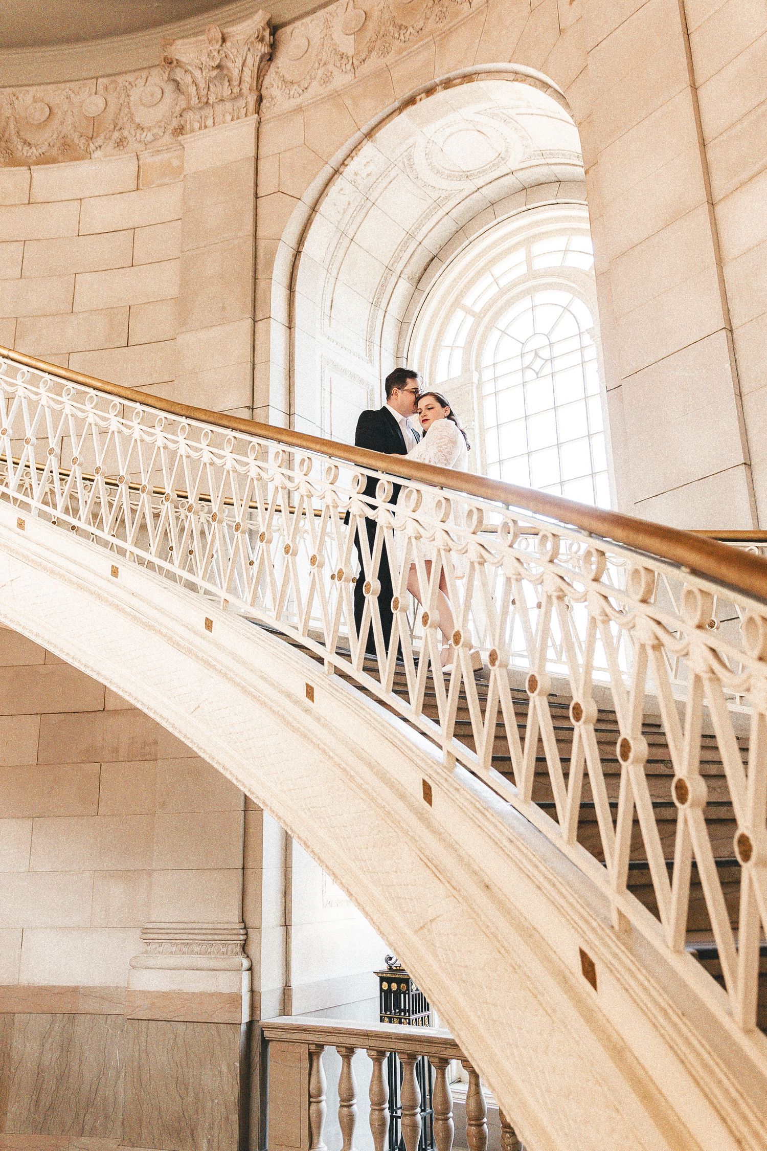 Bride and groom embrace on an ornate grand staircase with white iron railings in an elegant historic building.