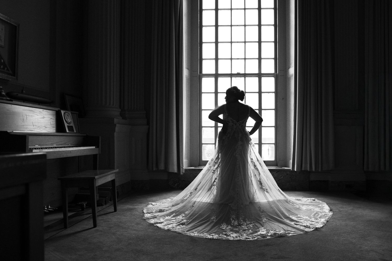 Bride in a long lace wedding gown silhouetted against tall window in black and white photo.