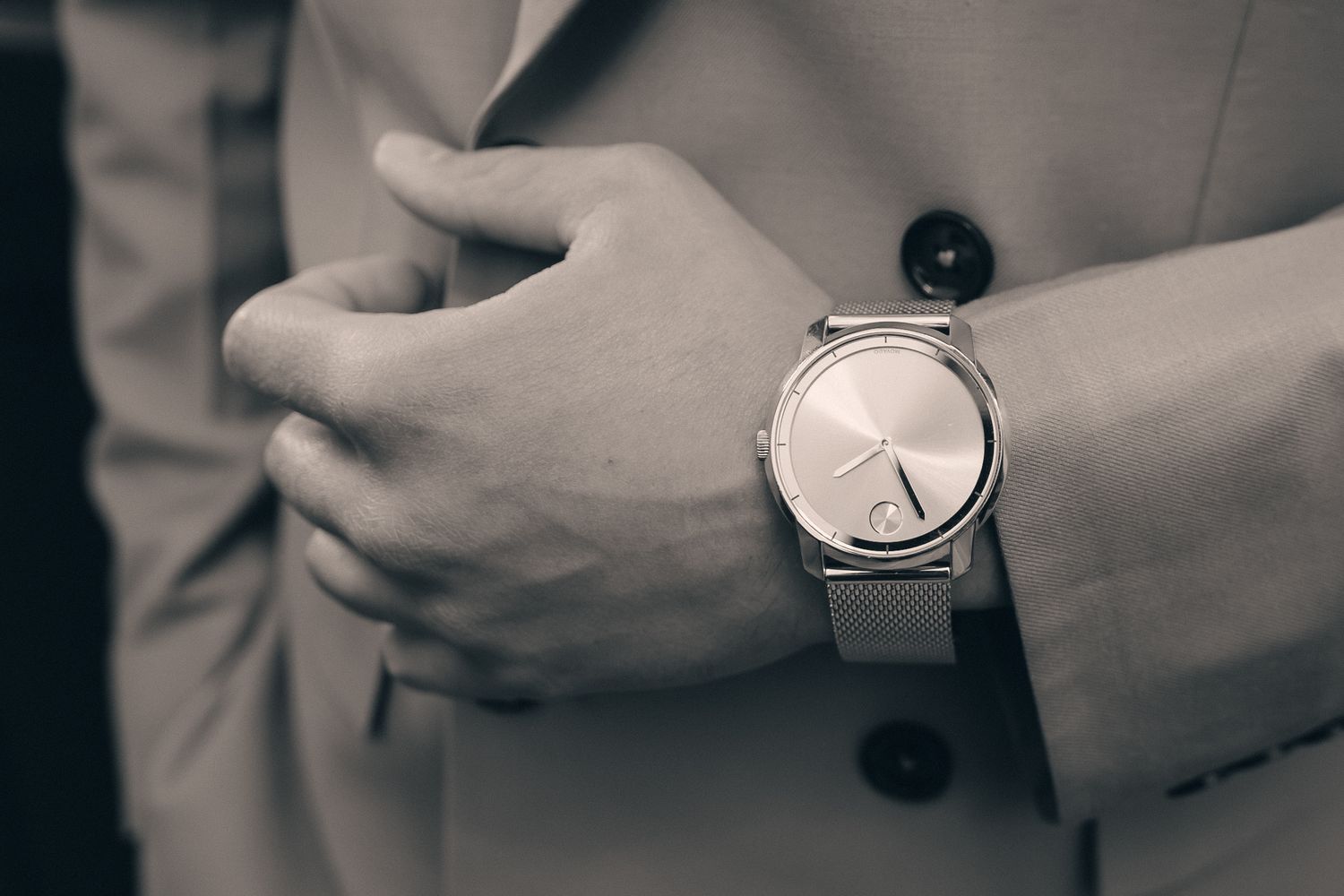Close-up of a man in a suit wearing a minimalist silver mesh-band wristwatch, captured in black and white.