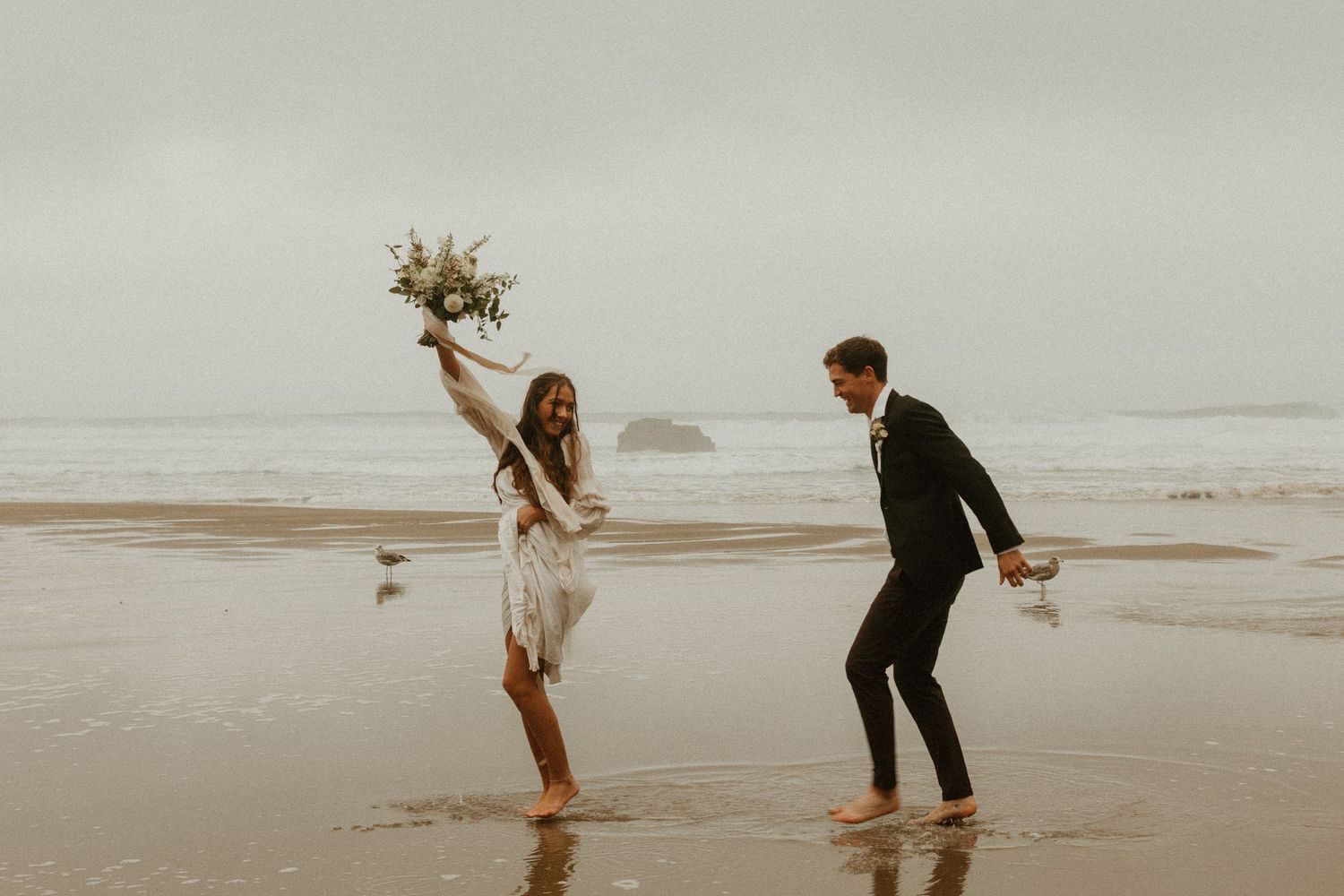 Joyful couple celebrating on a misty beach, bride holding bouquet aloft in a boho wedding look.
