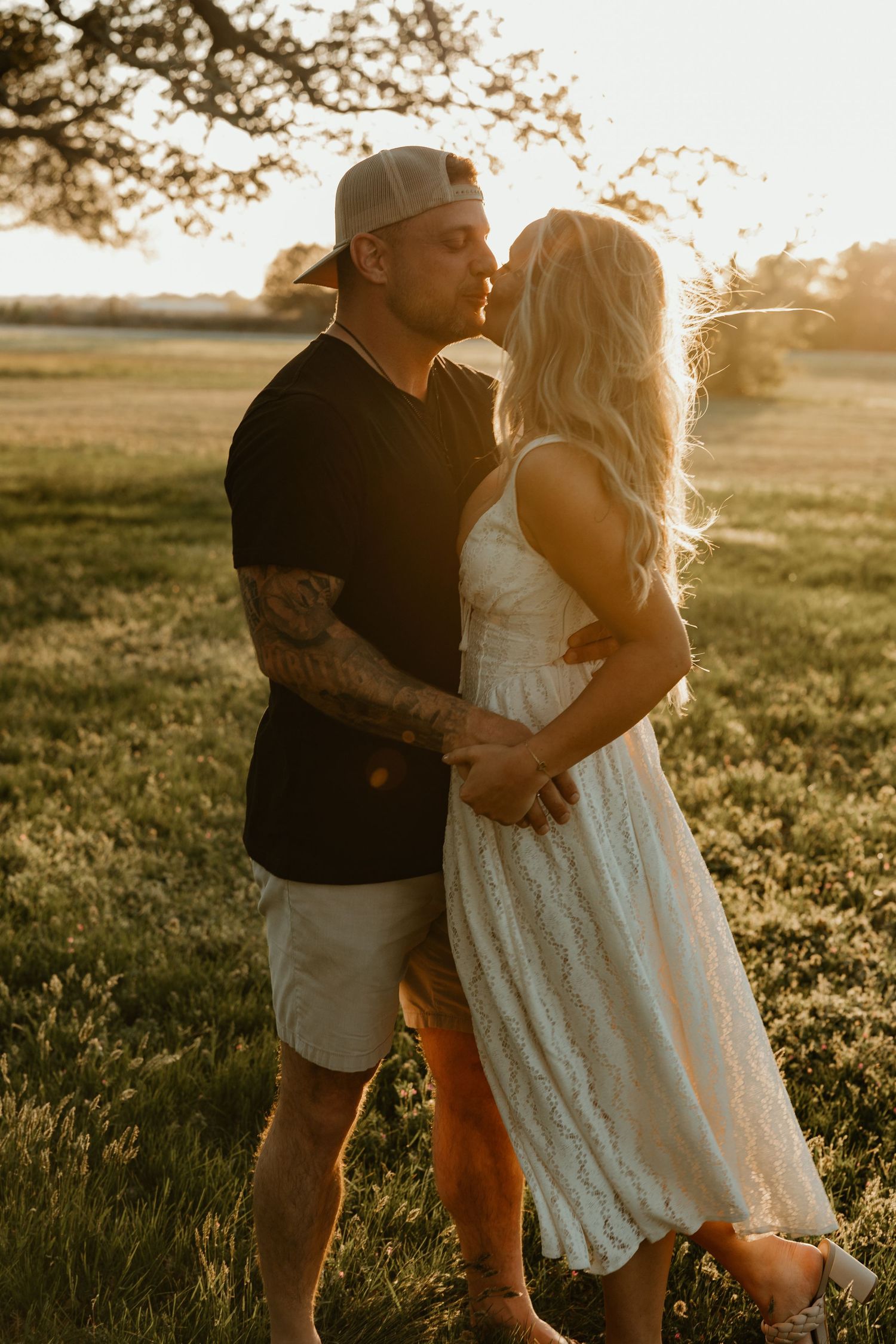 Tattooed man in black shirt kisses blonde woman in white lace dress at golden hour.