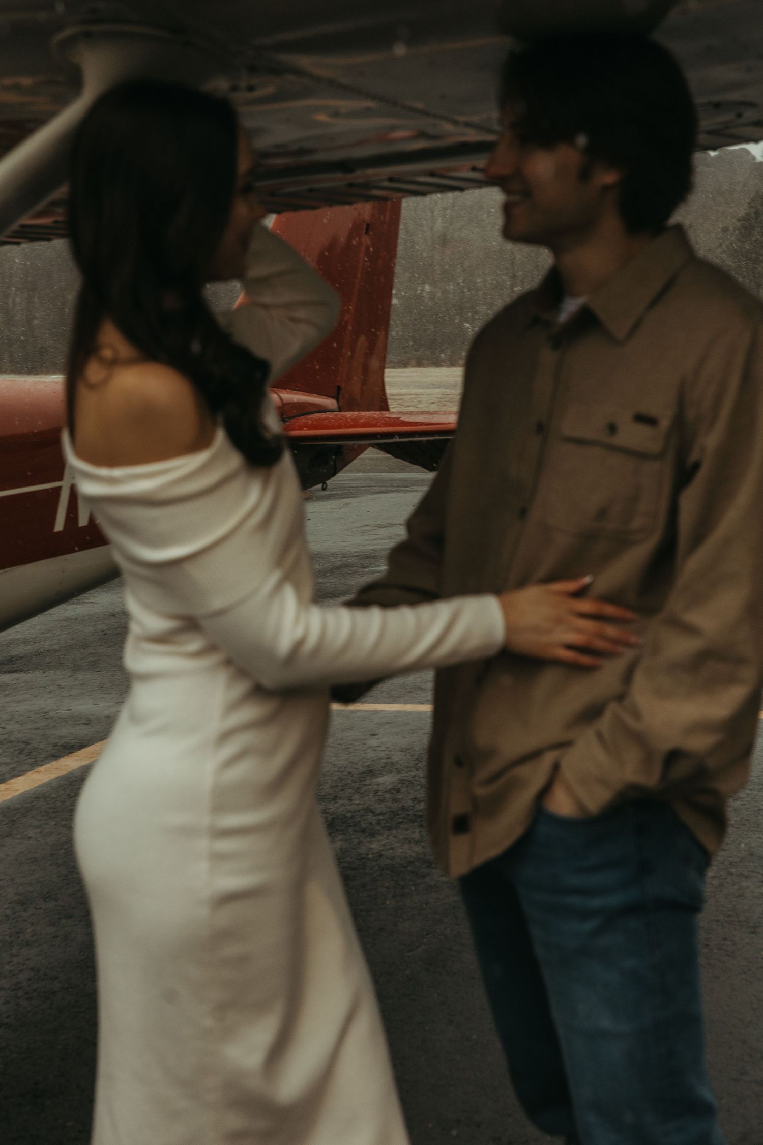 Couple holding each other beside a vintage red airplane on a rainy airstrip engagement shoot.