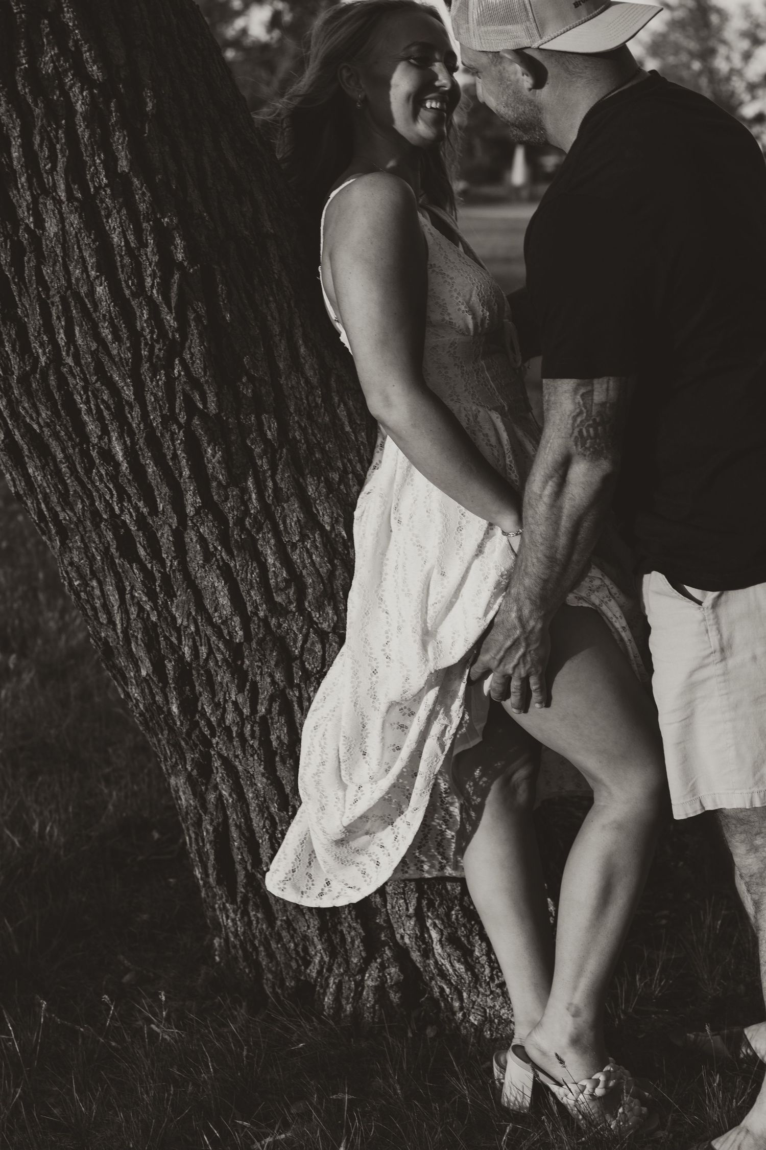 Black and white photo of a couple sharing a romantic moment leaning against a large tree outdoors.
