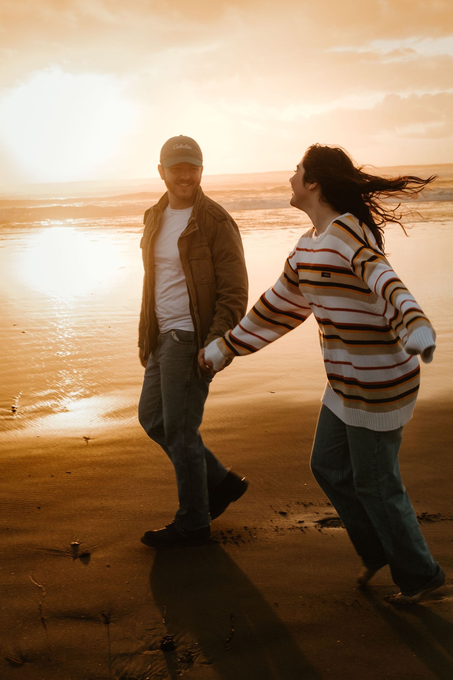 Couple holding hands and walking playfully on a sandy beach during a golden sunset.