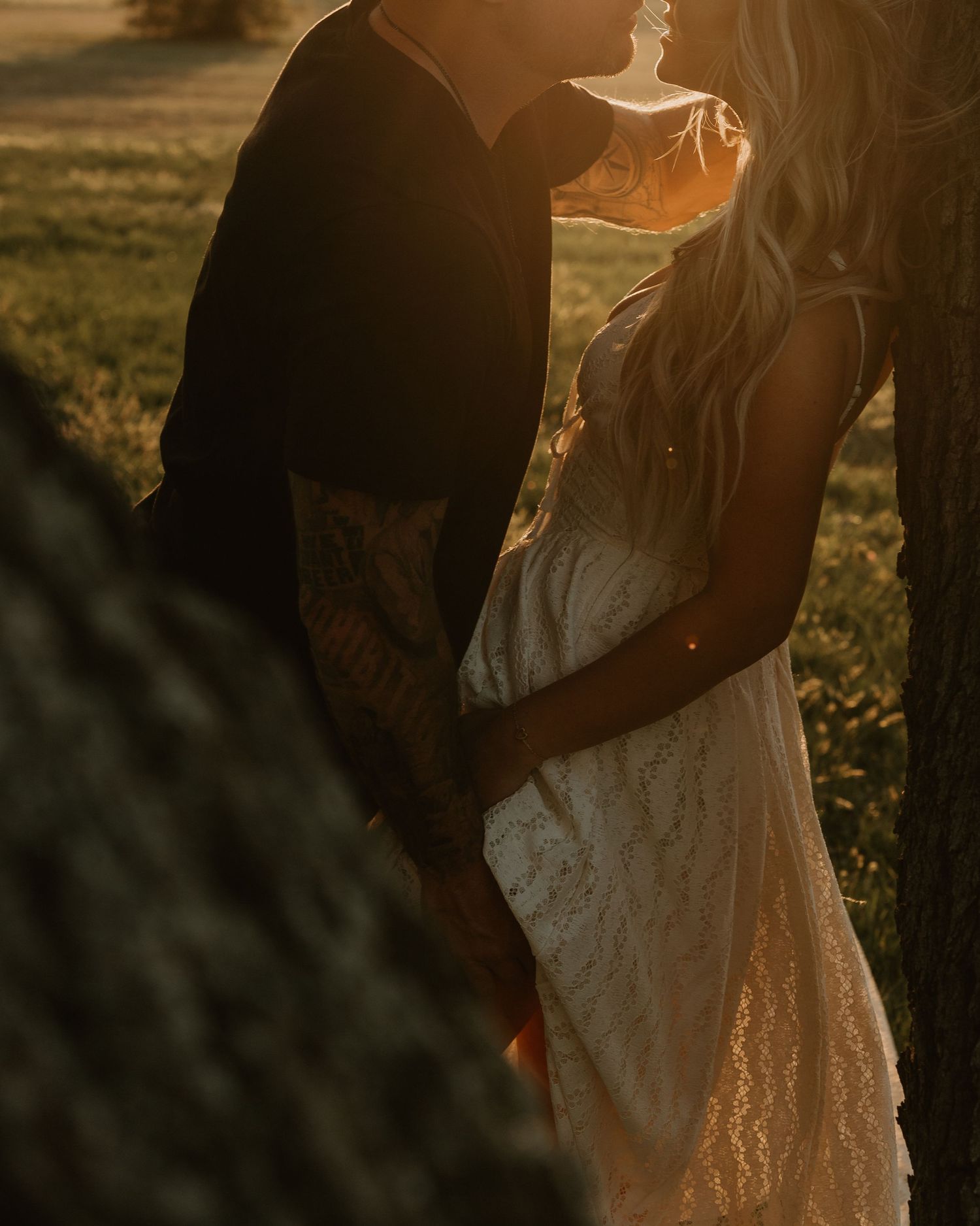 Romantic couple embracing at sunset, man with tattoos holding woman in lace dress by a tree.