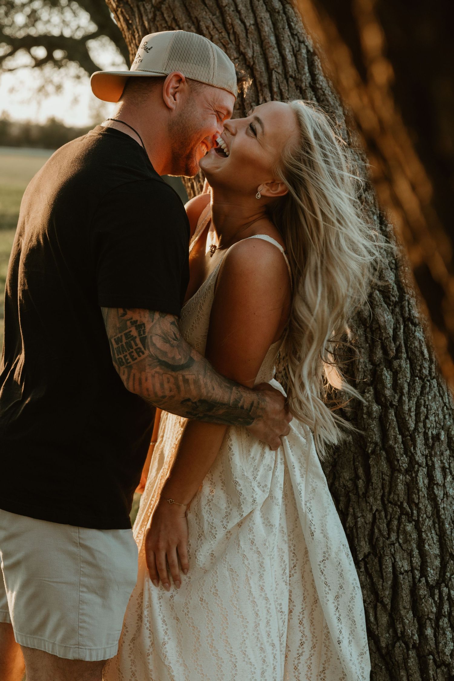 Laughing couple embracing against a large tree trunk in warm golden sunset light.