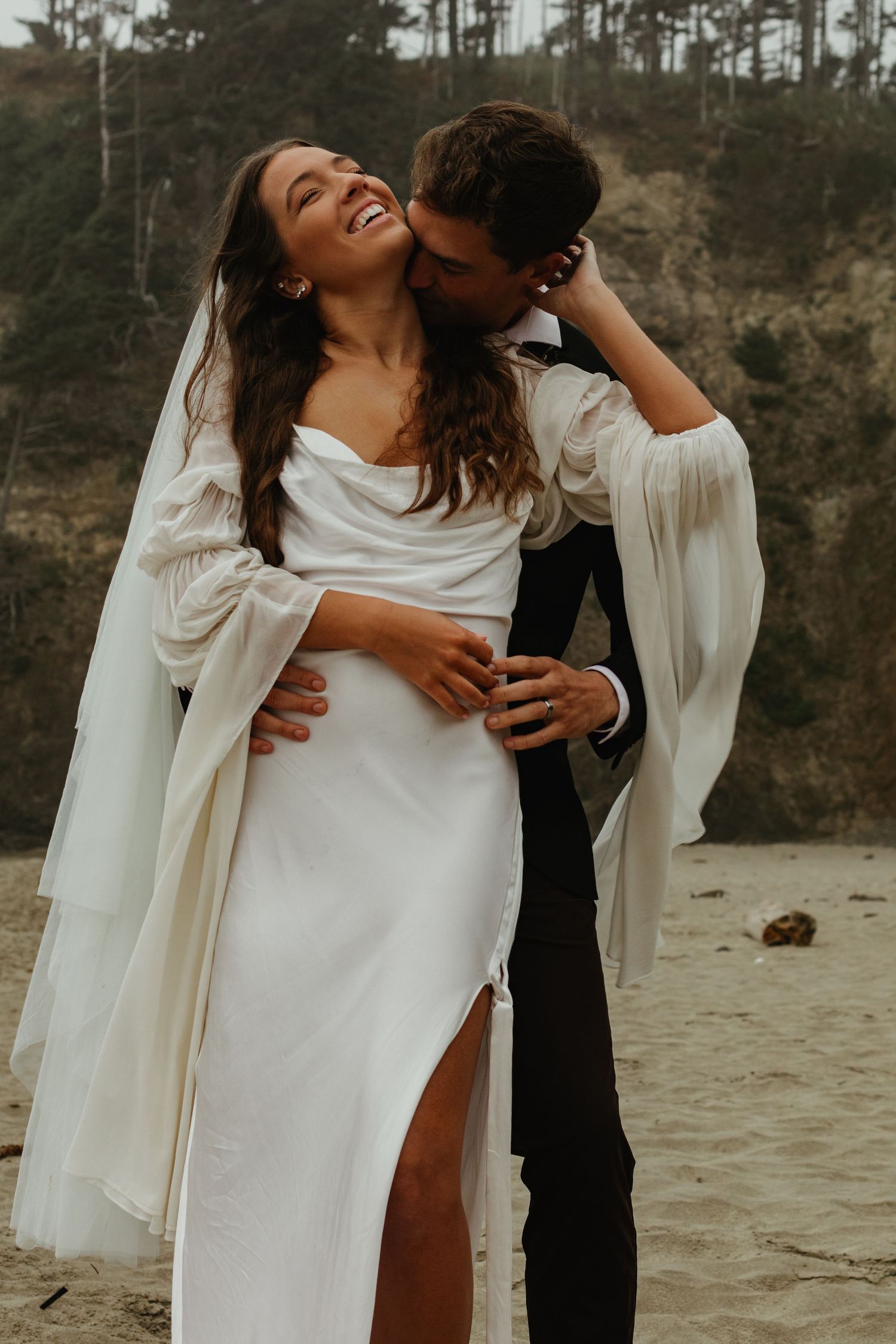 Joyful bride in flowing white gown laughs as groom kisses her neck on a sandy beach.