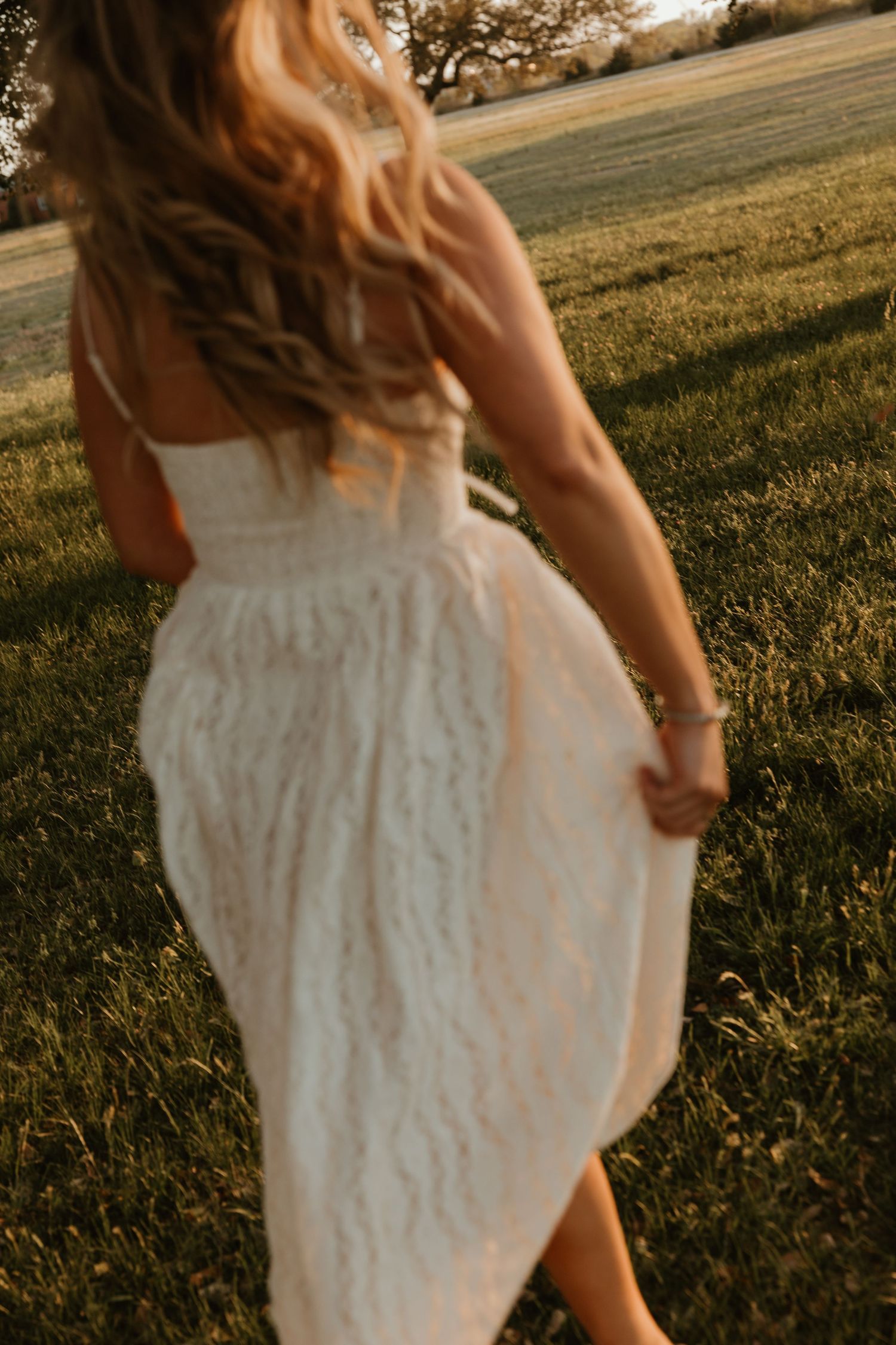 Back view of a blonde woman in a white lace dress walking through a green field at golden hour sunset.