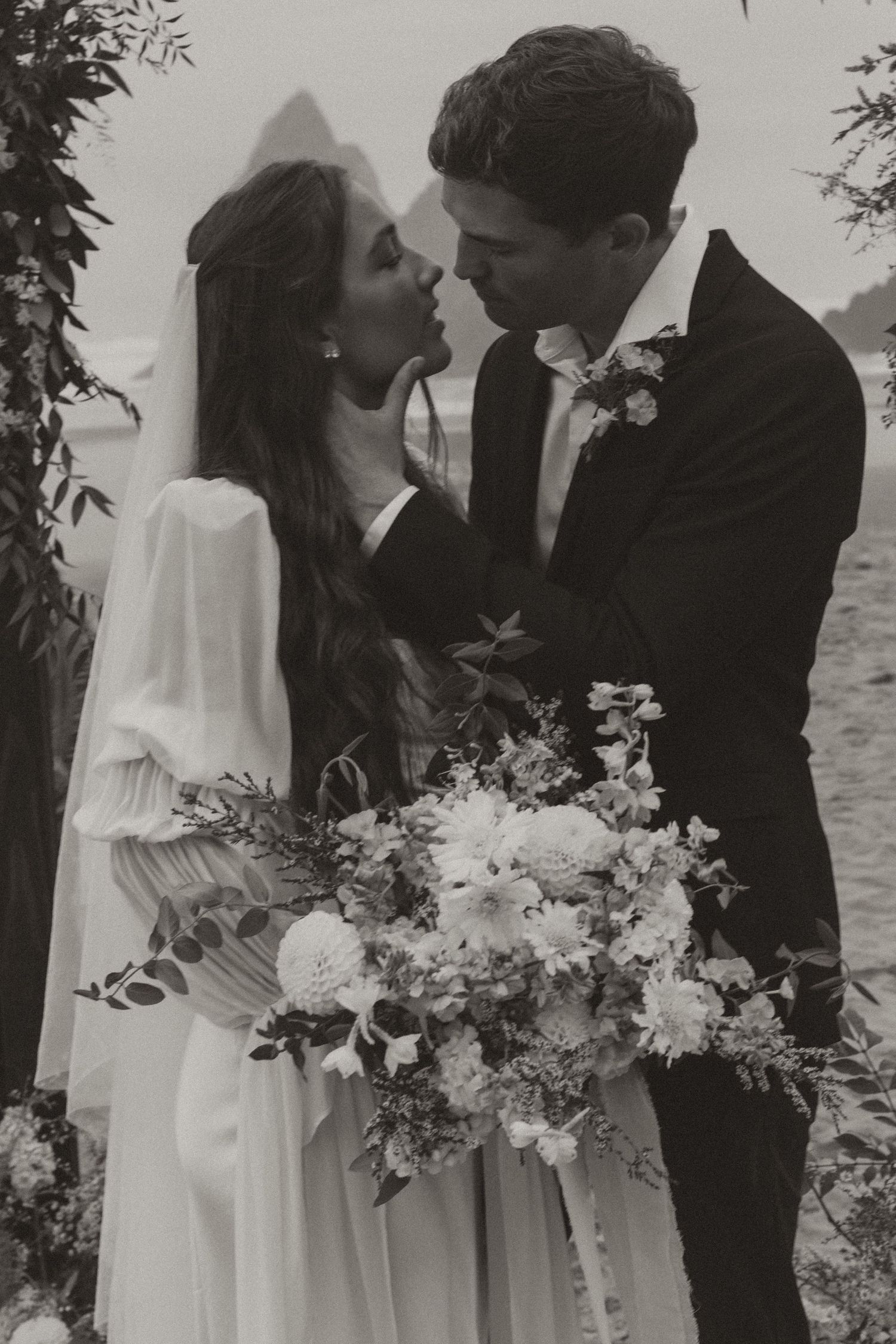 Black and white photo of bride and groom nearly kissing beneath a floral arch.