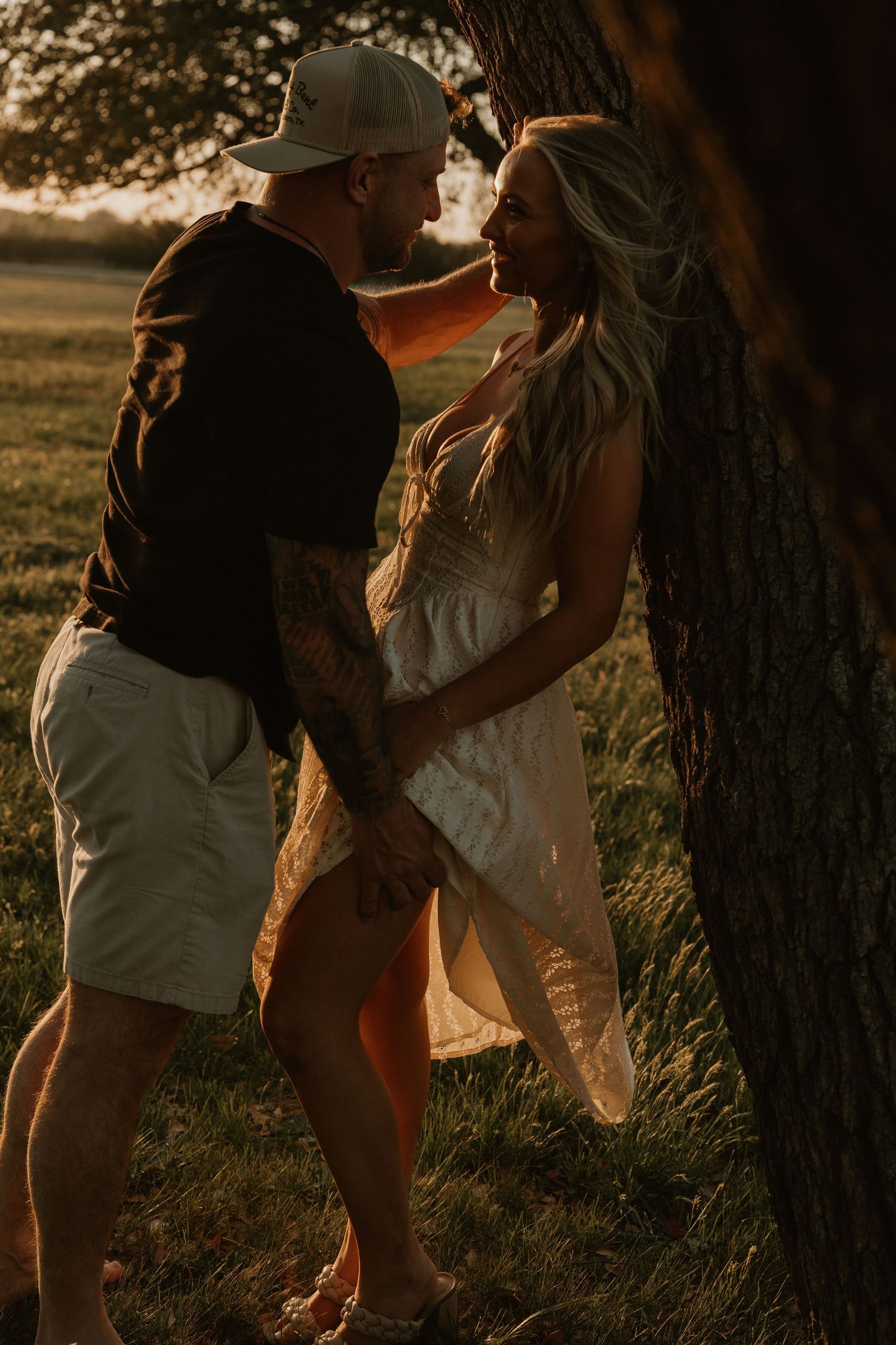 Couple sharing a tender moment at golden hour beside a large tree in an open field.